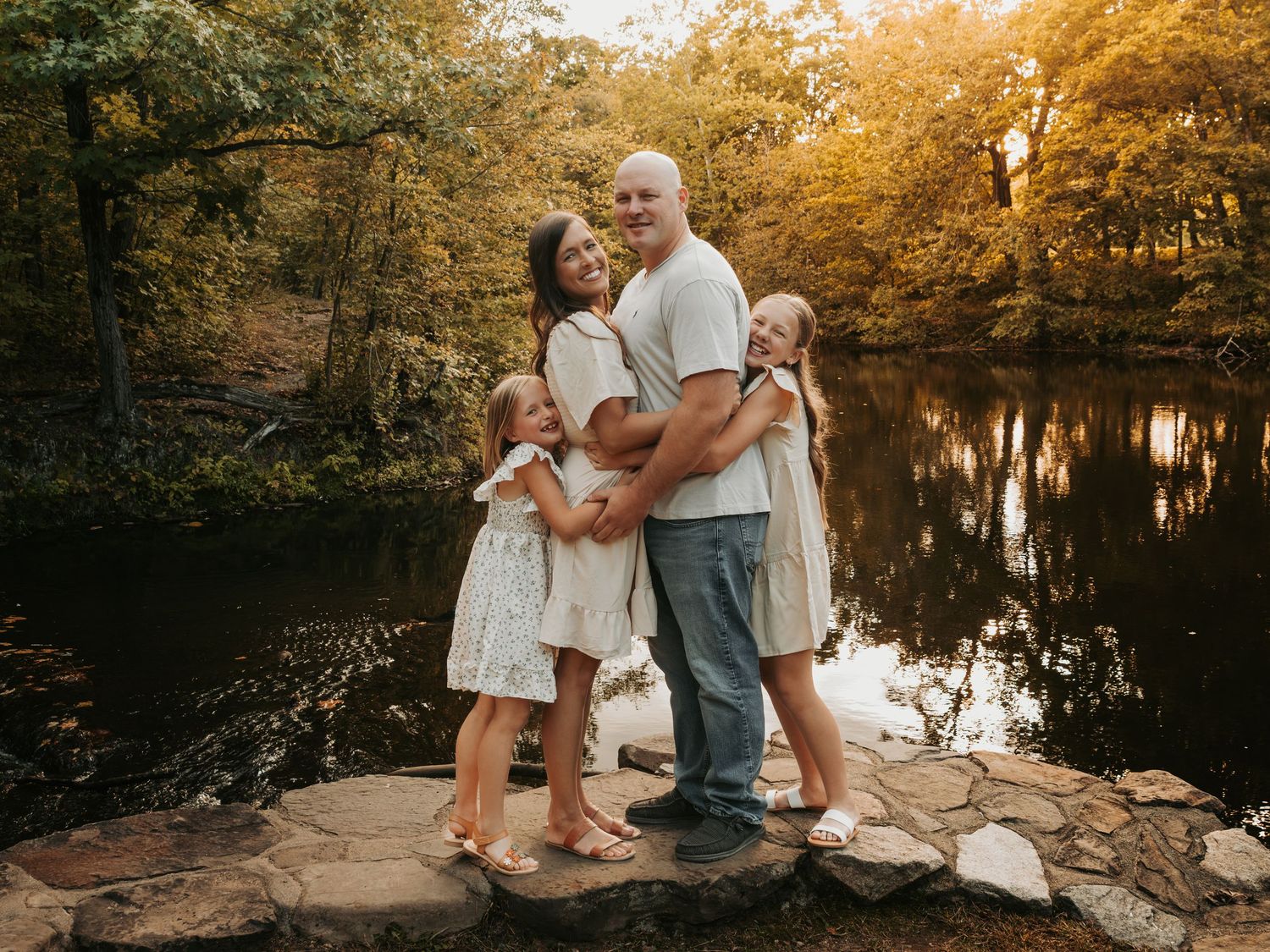Family of four posing on rocks by a reflective pond surrounded by autumn foliage in a warm golden light.