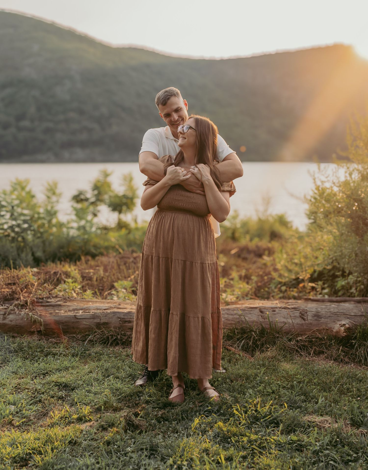 Couple embracing at golden hour by a lake with mountain backdrop, woman in rust tiered maxi dress.