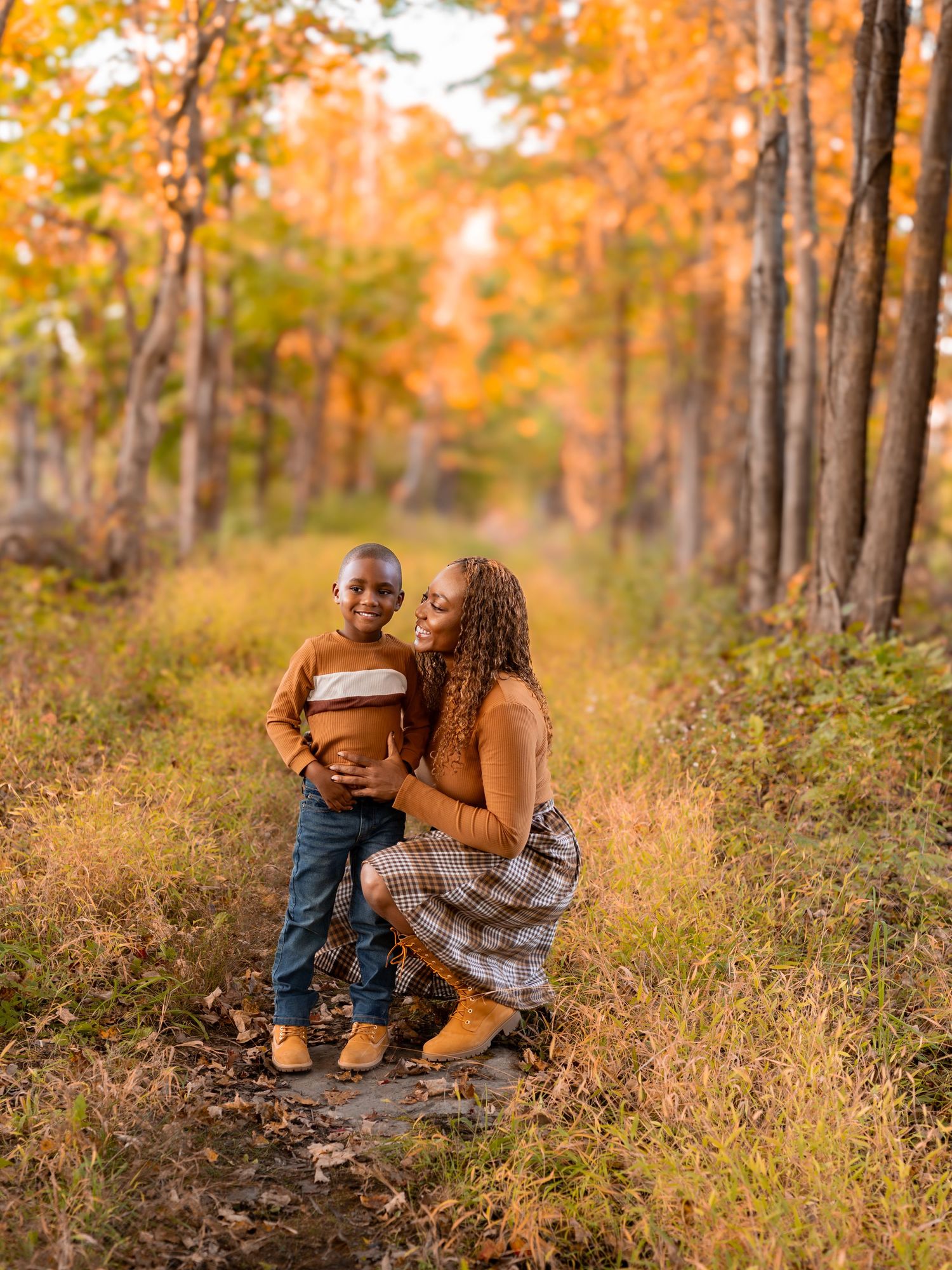 A mother kneels on a forest path hugging her young son amid vibrant autumn foliage in warm orange and gold tones.