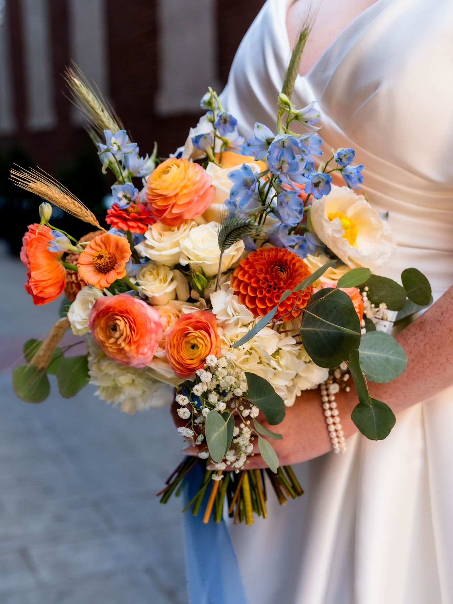 Bride holding a vibrant wedding bouquet with orange dahlias, blue delphiniums, cream roses, and eucalyptus foliage.