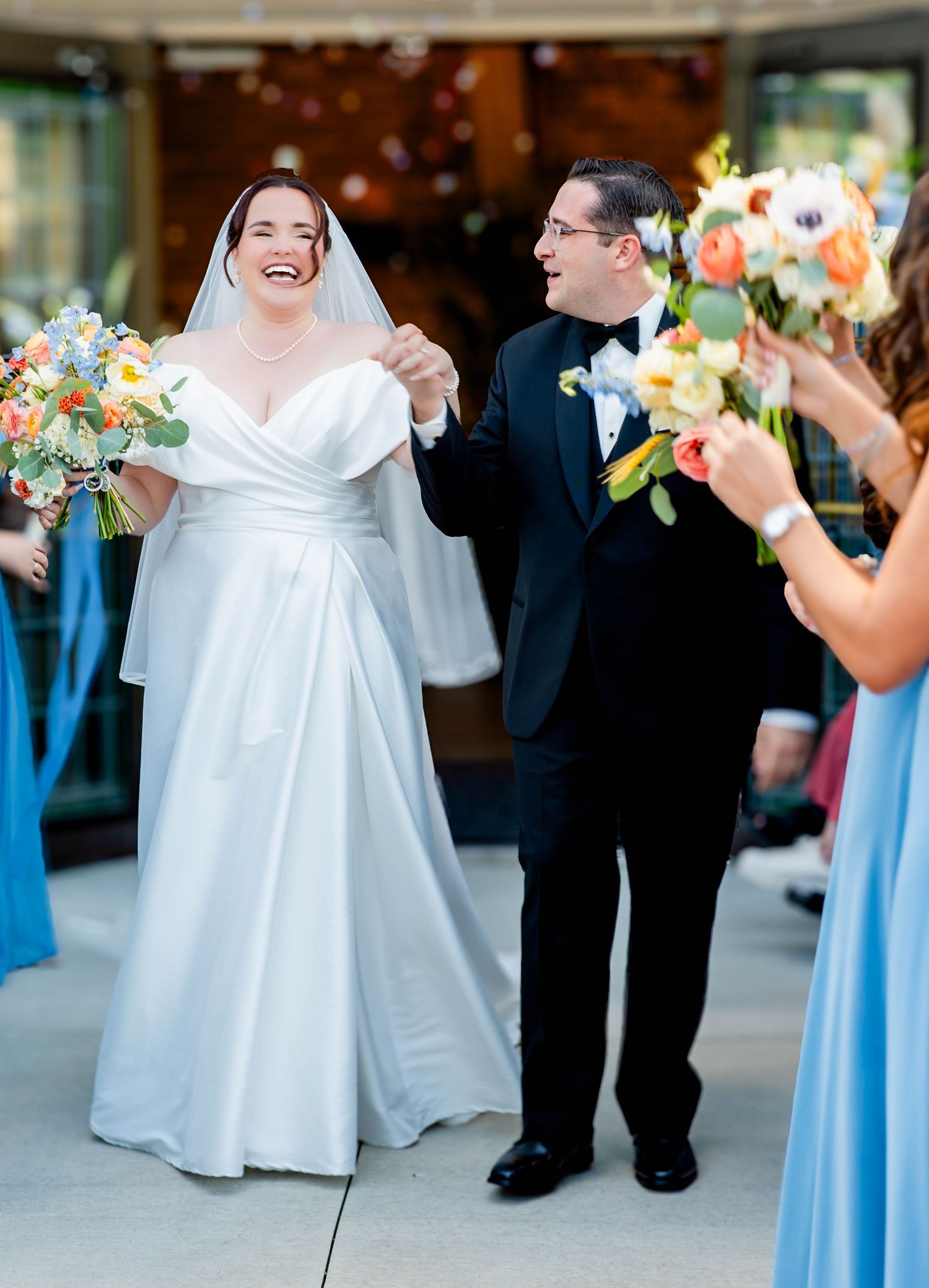 Joyful bride in white gown and groom in tuxedo walk down aisle, surrounded by guests holding colorful bouquets.