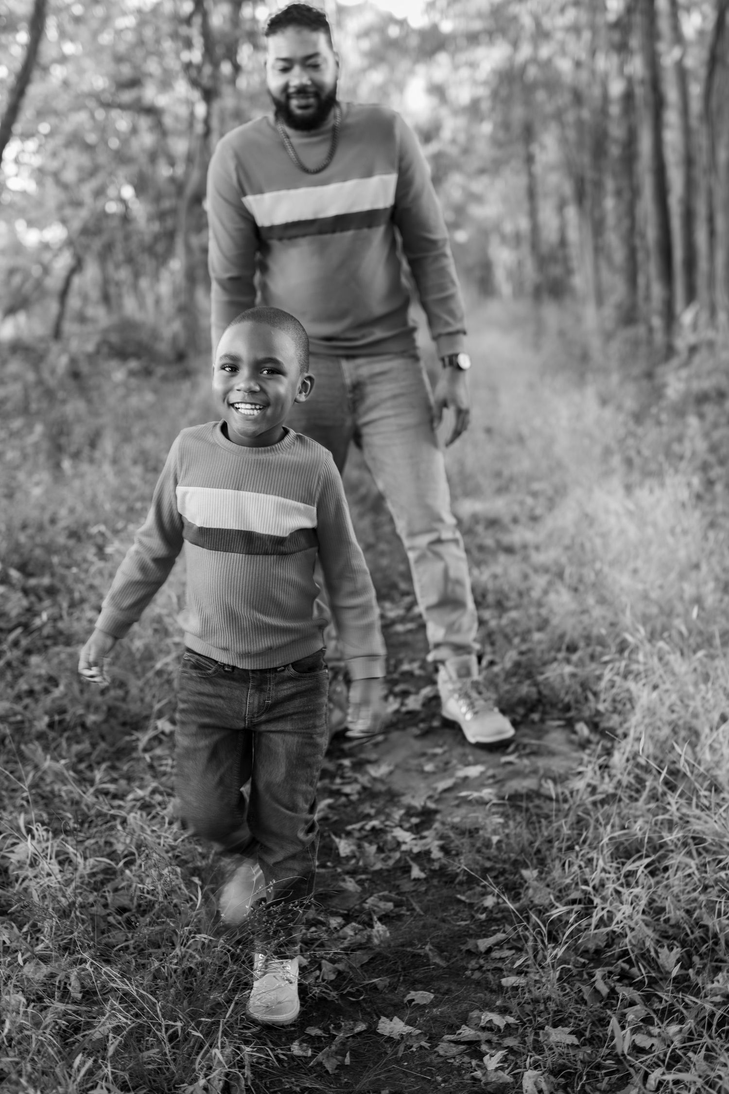 Black and white photo of a smiling boy and man in matching striped sweaters walking on a forest path.