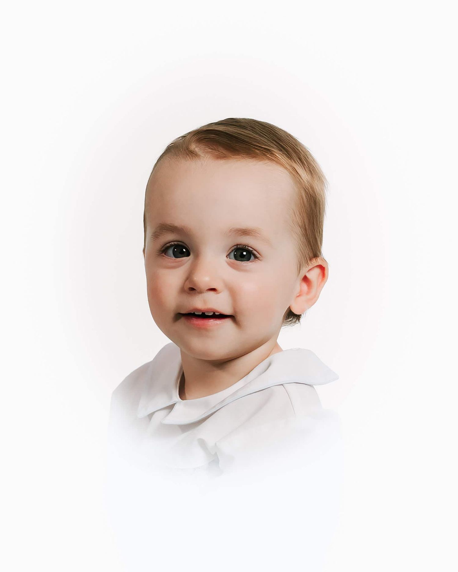 heirloom Portrait of a smiling toddler with light brown hair wearing a white shirt against a white background.