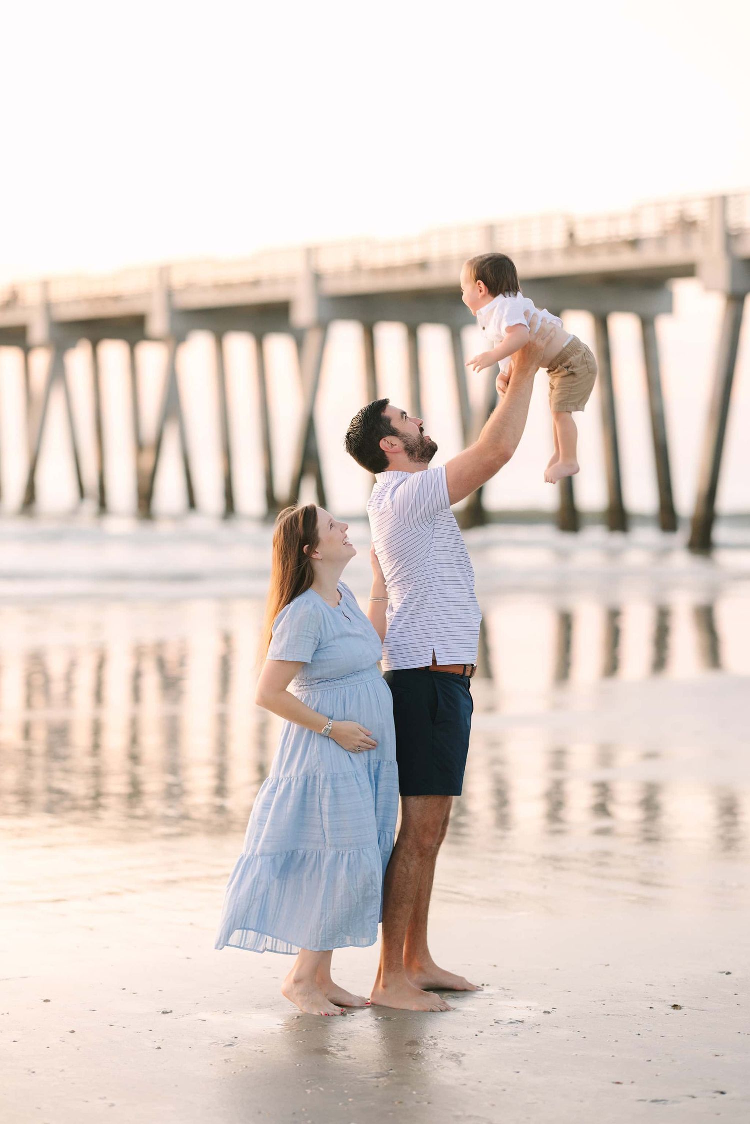 pregnant mom holding belly and laughing as her husband tosses toddler boy into the air in front of the Jacksonville beach pier