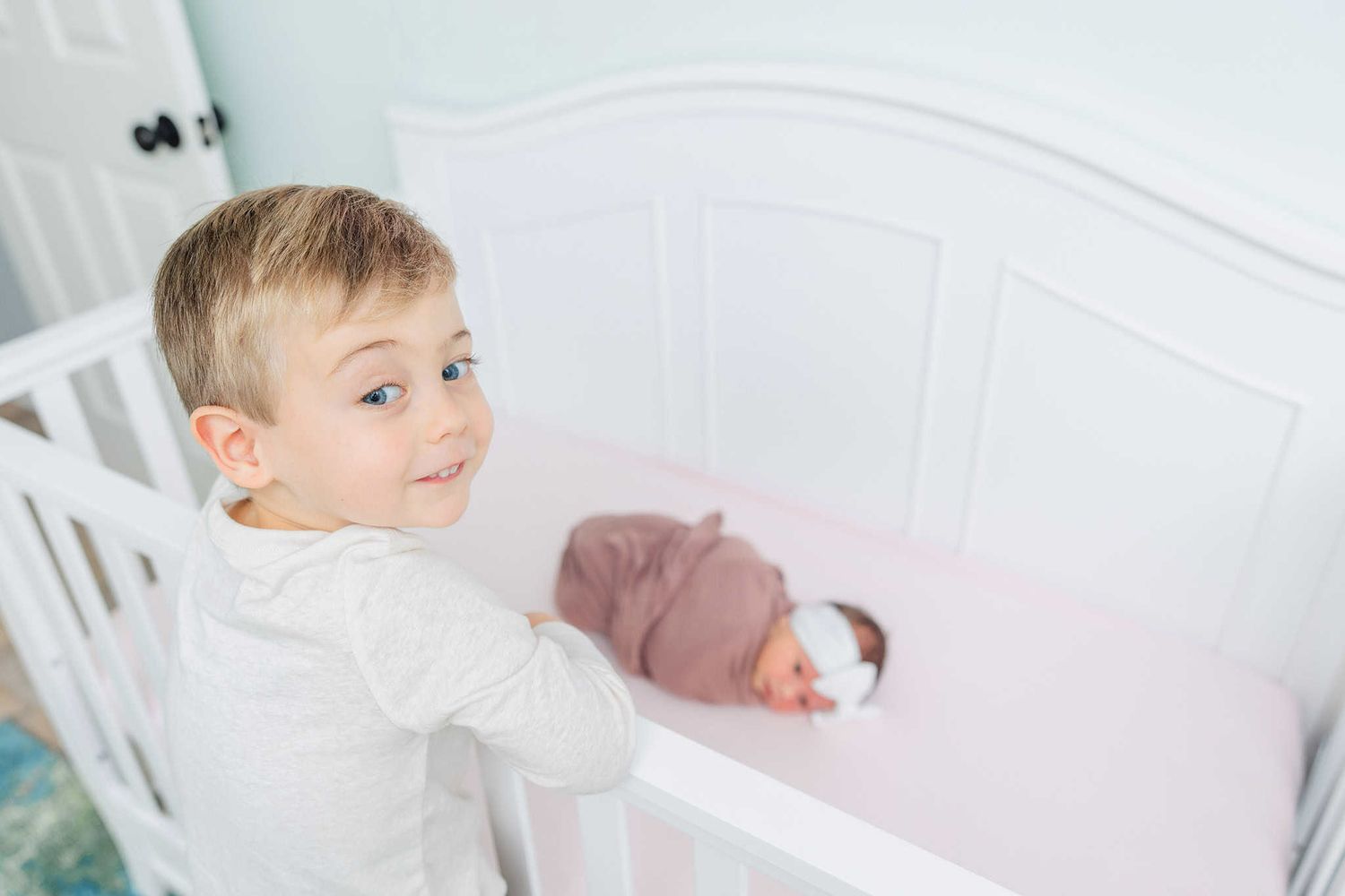 older brother standing on the crib looking at camera while newborn baby sister sleeps in the crib