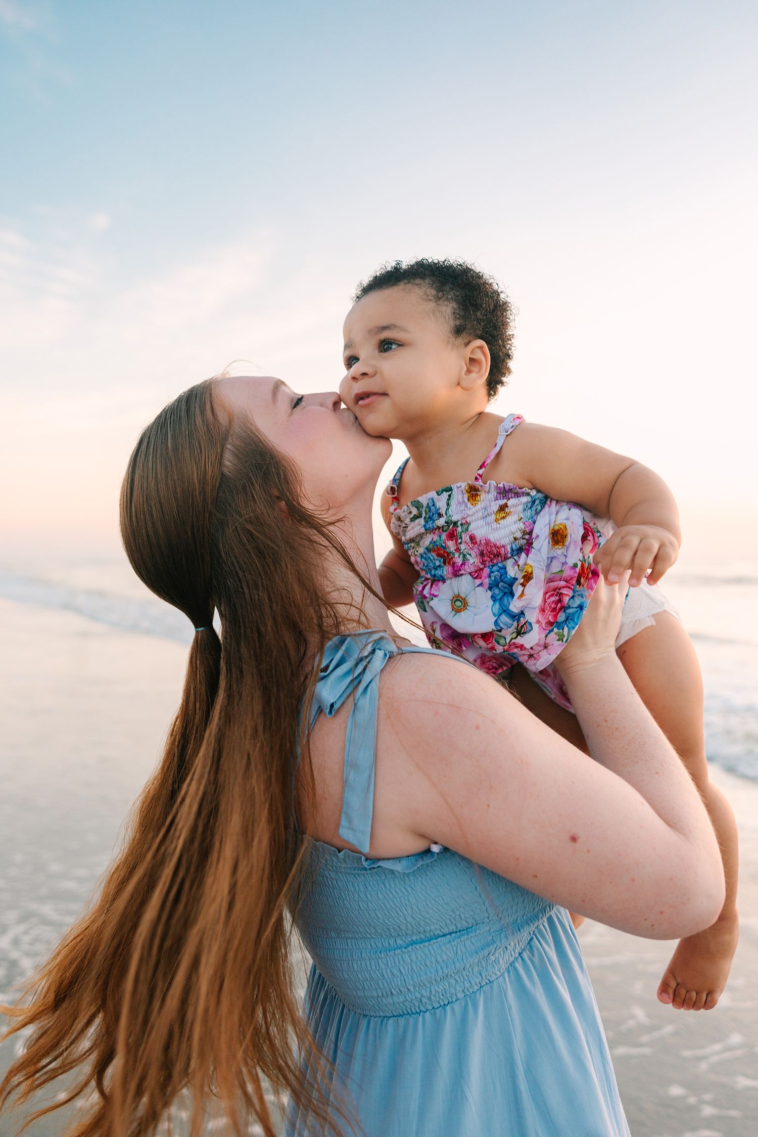 mom holding baby girl up in the air giving her a kiss on the cheek at Jacksonville beach, fl sunrise