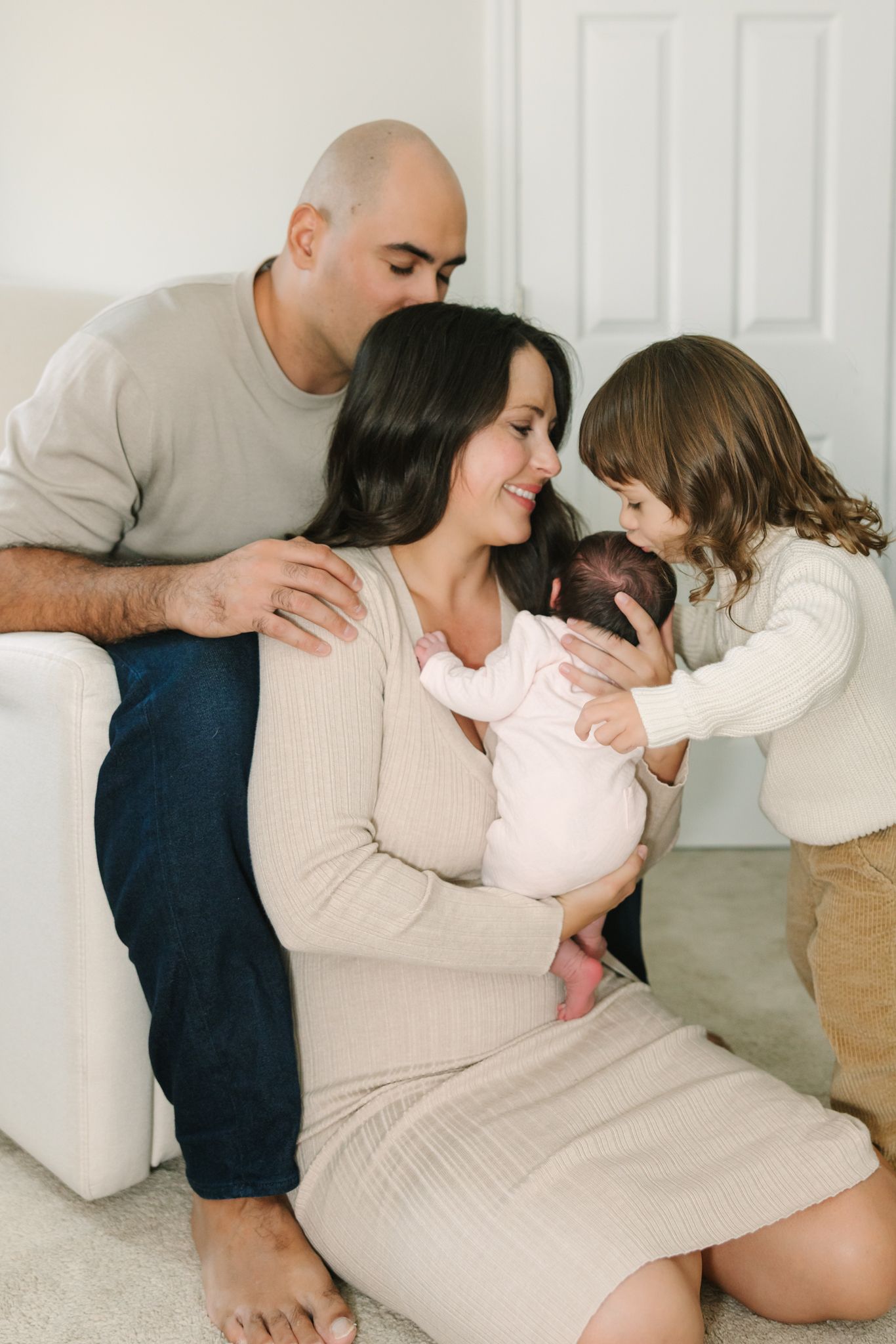 mom sitting on the floor holding baby while toddler kisses baby's head. dad sitting on chair behind mom kissing her