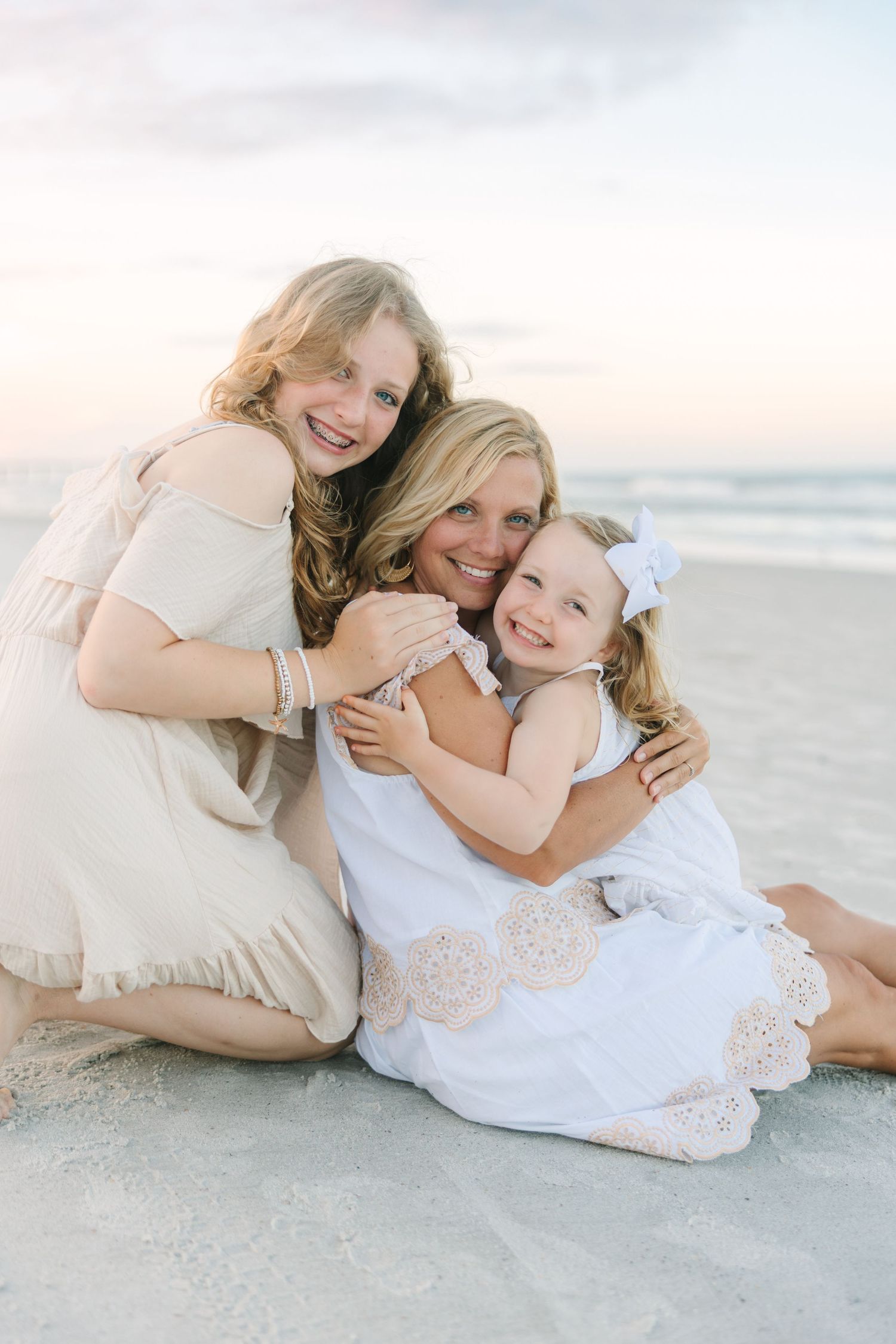 mom snugglinh with both daughters on the beach wearing white