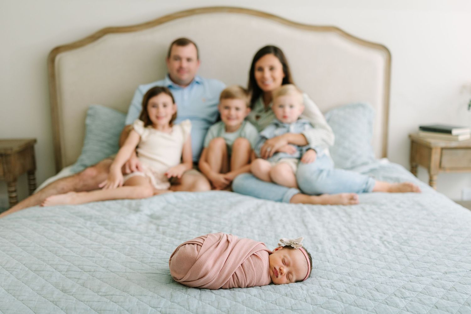 Newborn baby swaddled in pink on bed with family of six smiling behind in a bright bedroom