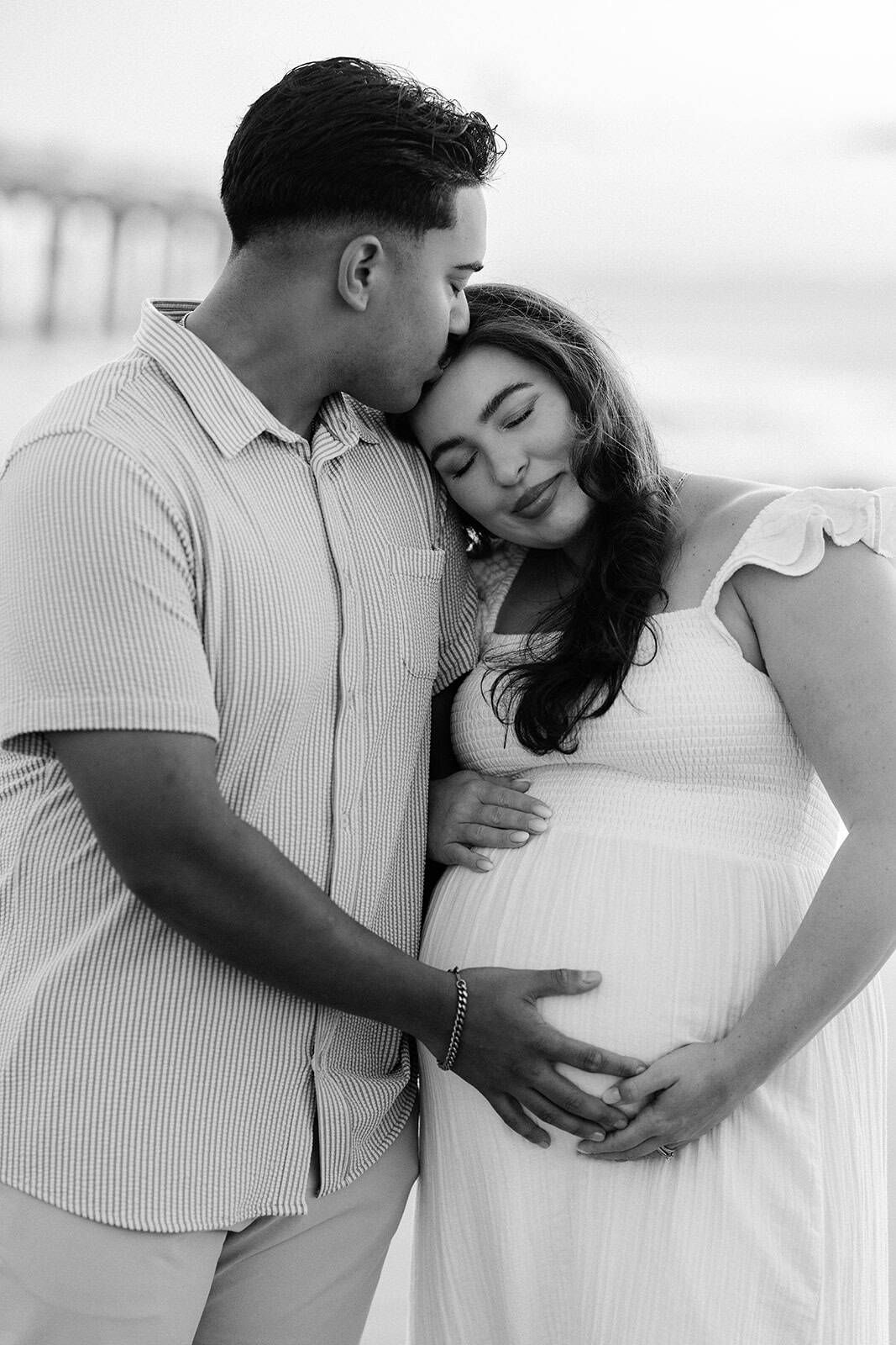 black and white image of dad holding and kissing pregnant wife at Jacksonville beach pier