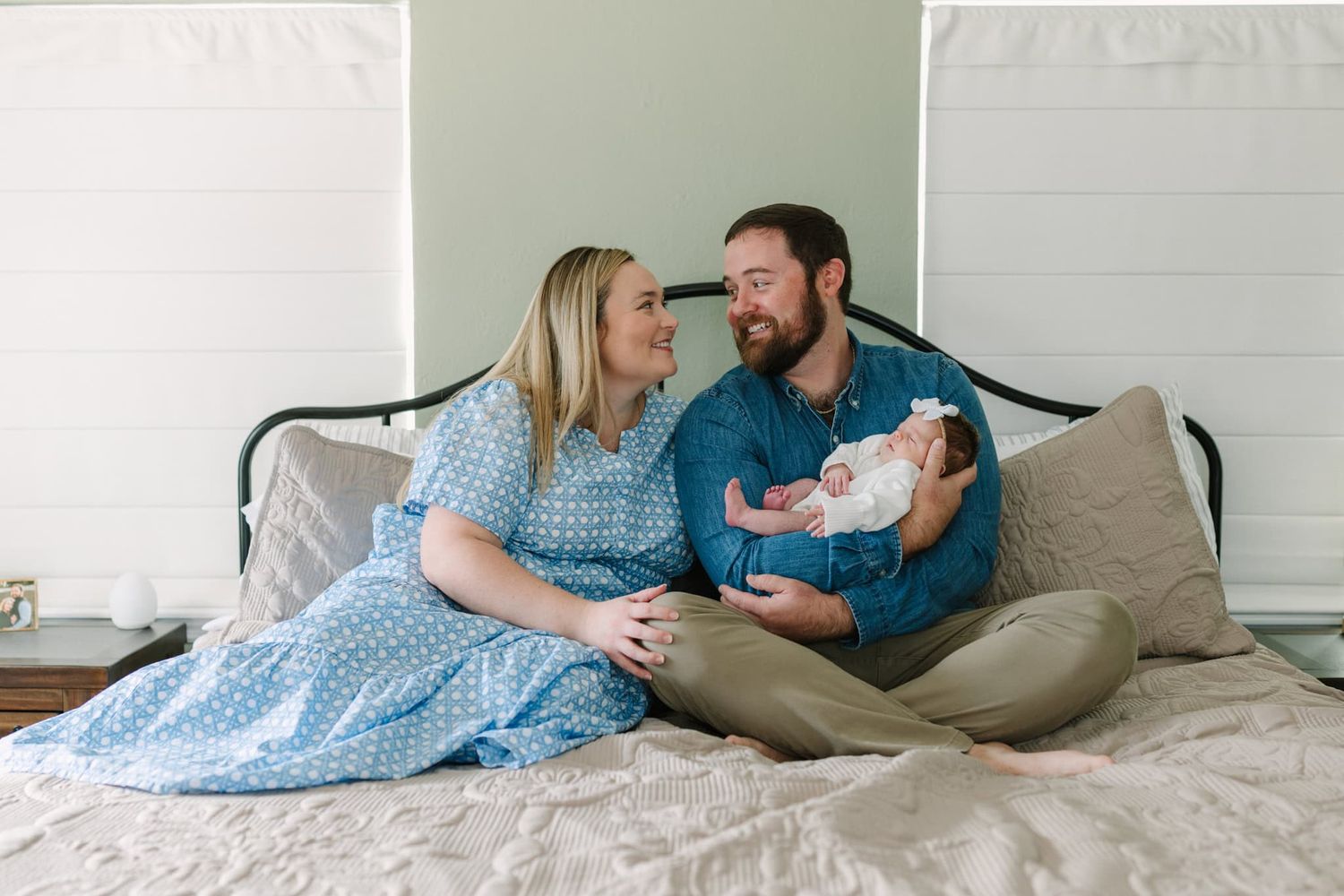 mom and dad wearing blue and sitting on their bed looking at each other and smiling while holding newborn baby girl