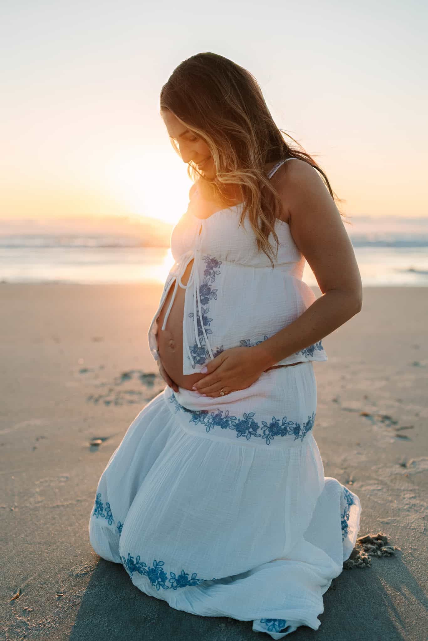 Pregnant woman in white dress kneeling on beach at golden sunrise in Ponte vedra beach fl, cradling her baby bump.