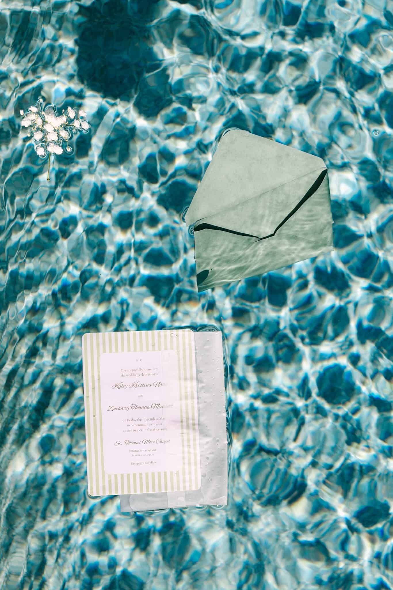 Elegant wedding invitation suite with sage green envelope floating on shimmering turquoise pool water.