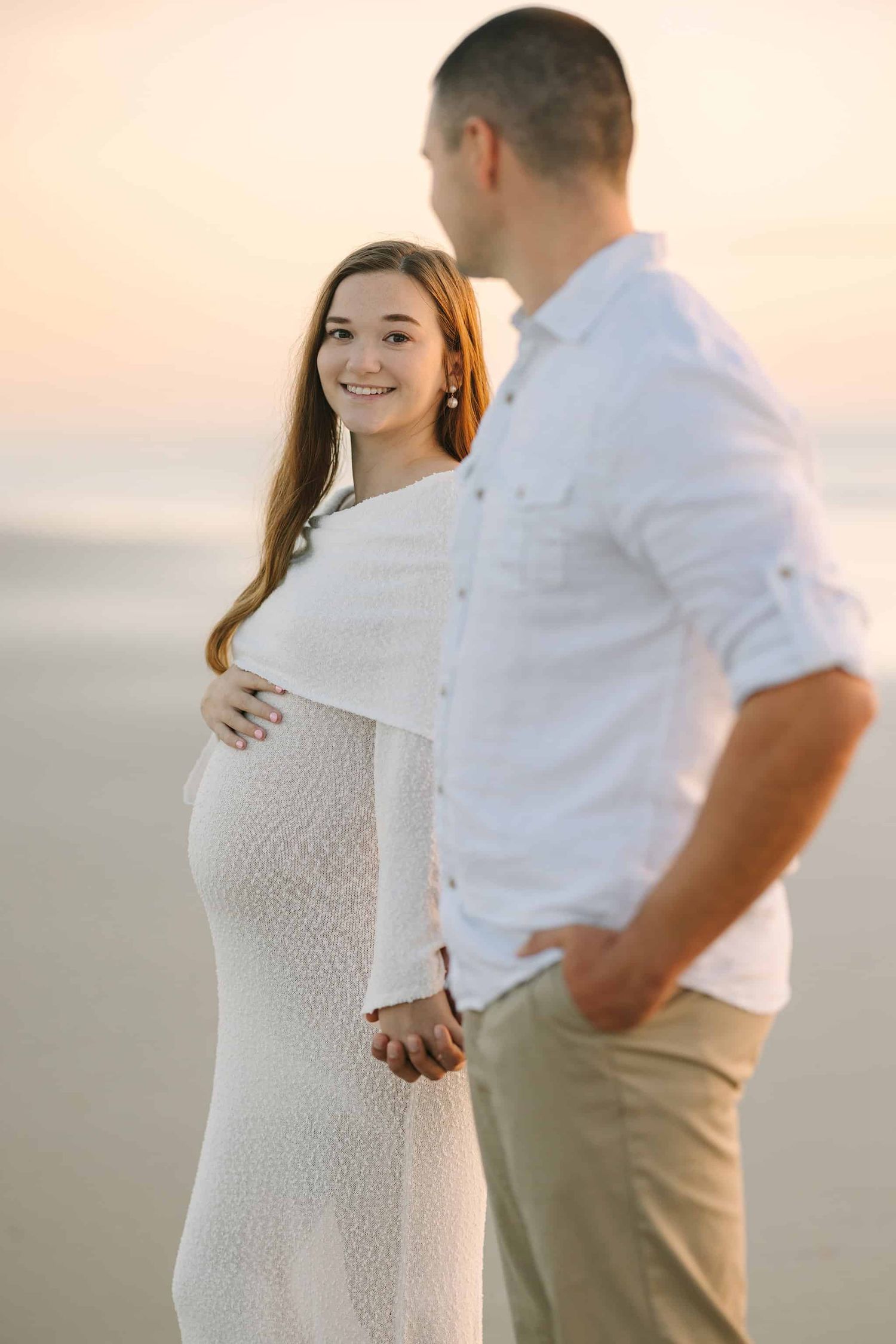 pregnant mom in white dress on the beach, looking at her past her husband who looks back at her