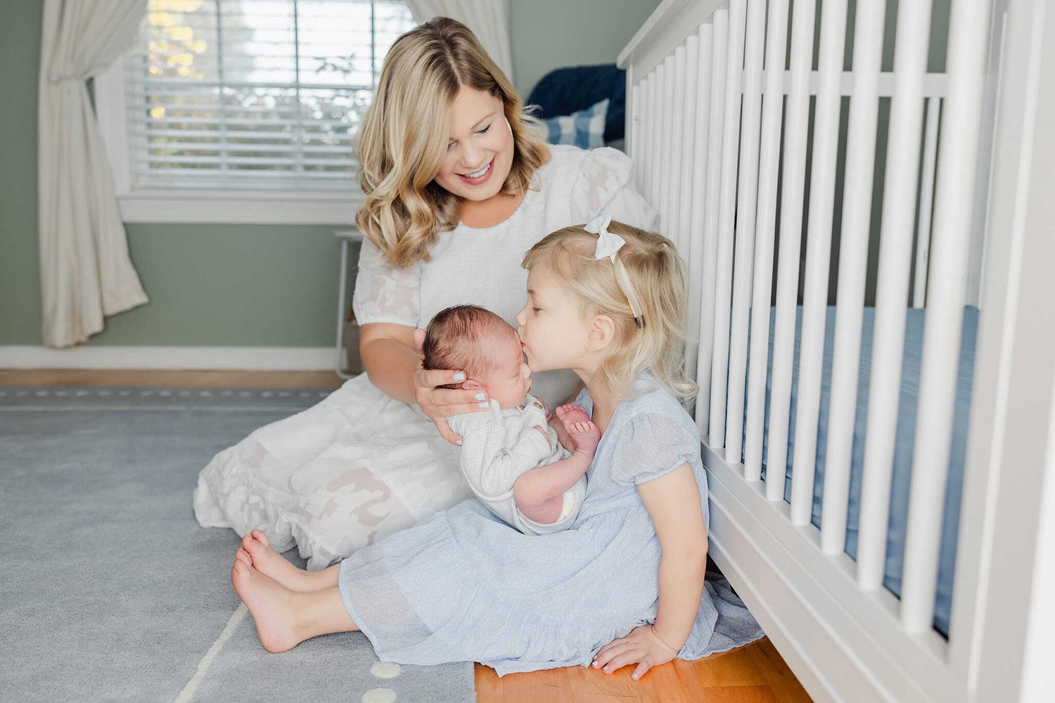 big sister holding and giving newborn brother a kiss while mom sits and helps her hold him