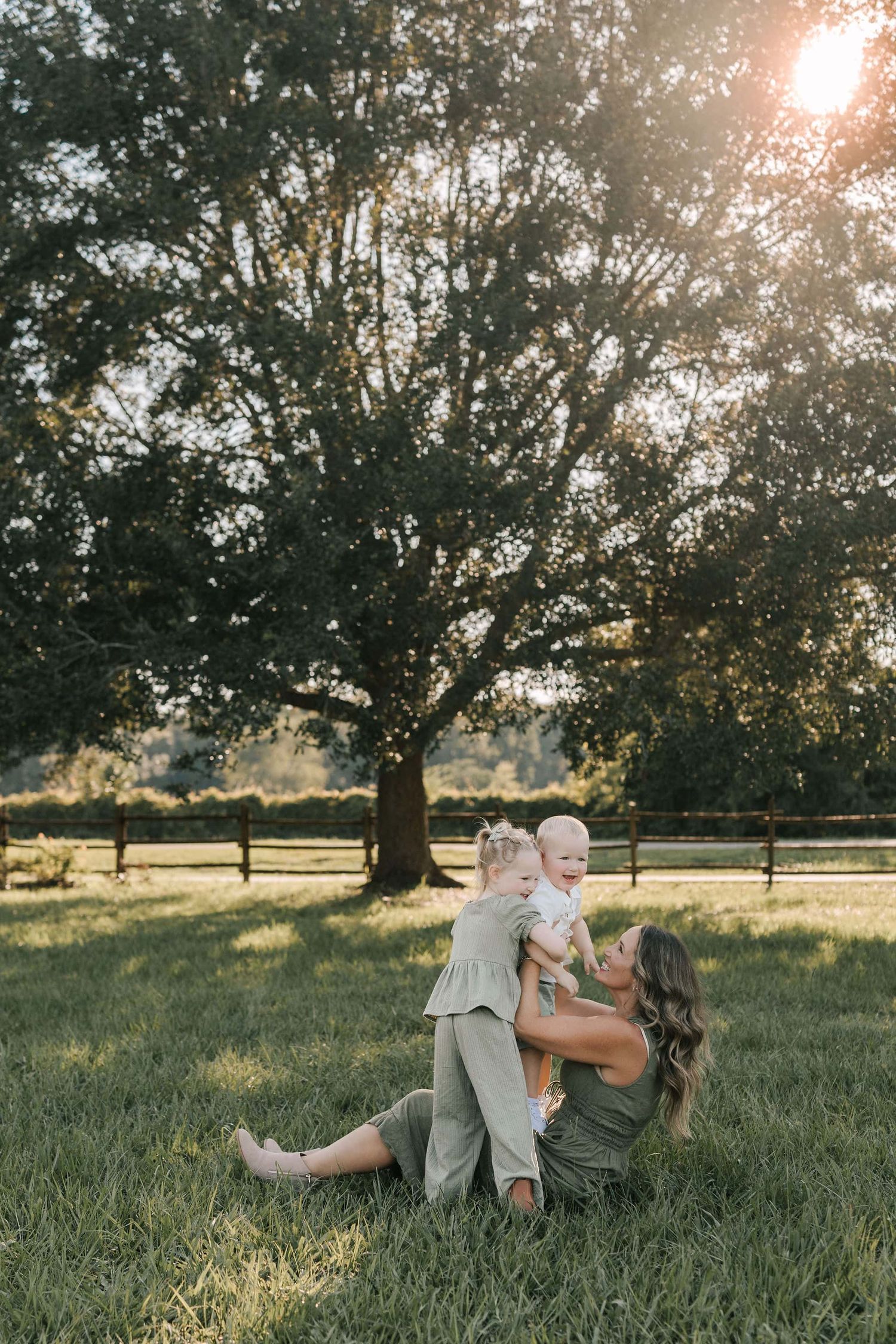 A mother sits on lush green grass holding two young children, bathed in golden sunlight beneath a large oak tree