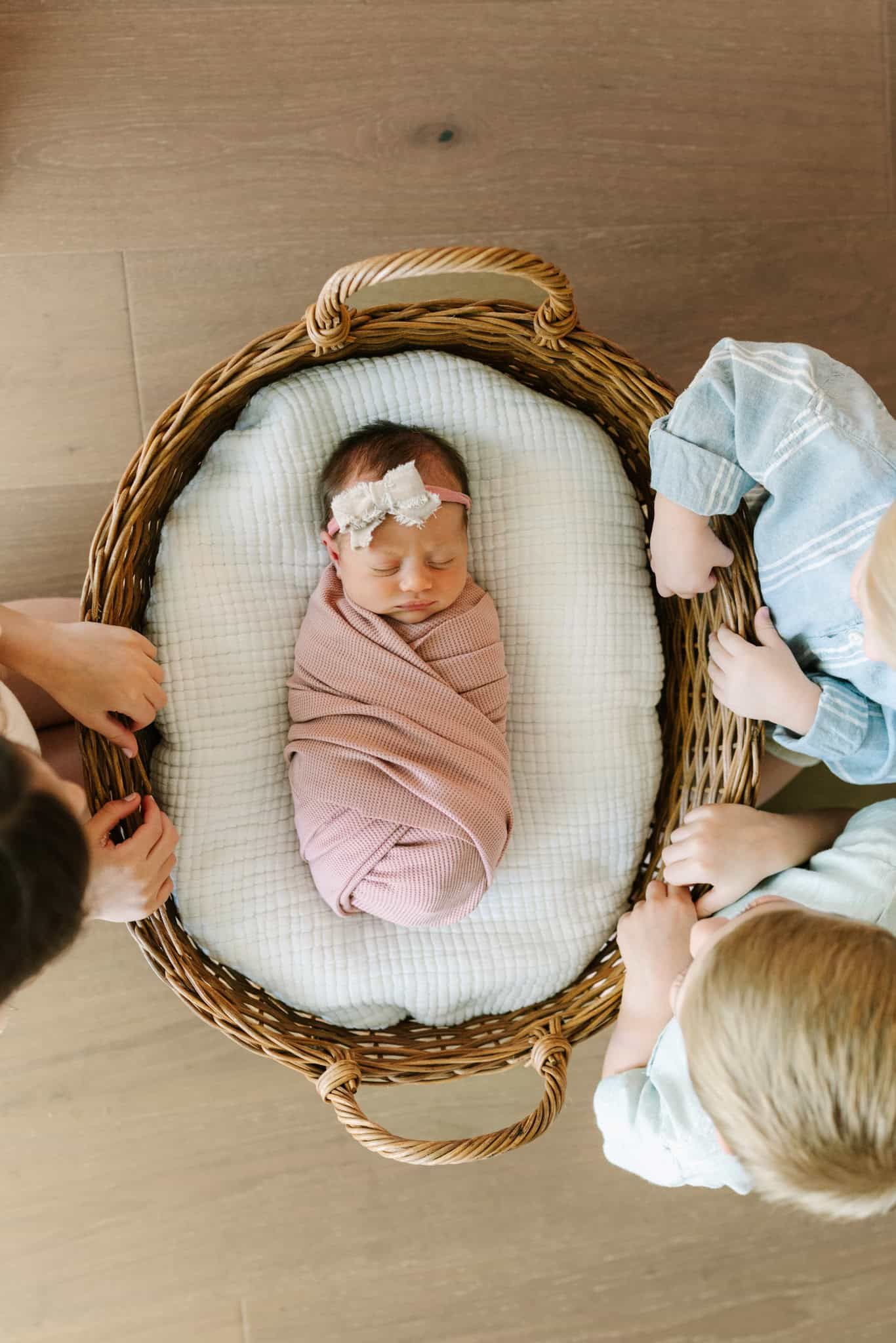 Newborn baby girl swaddled in pink in a wicker basket, viewed from above with three siblings looking on.