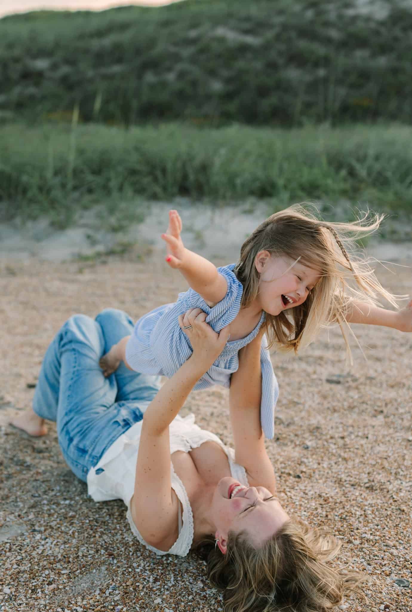 A mother lies on sandy Ponte Vedra beach lifting her laughing young daughter in the air, both joyful with green dunes behind.