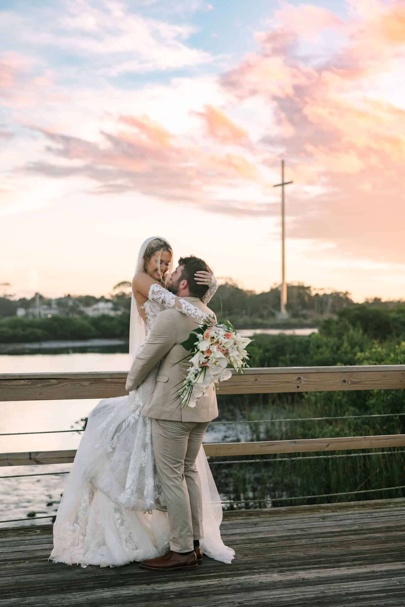 groom holding bride up while they look at each other adoringly with st Augustine cross in the background during sunset