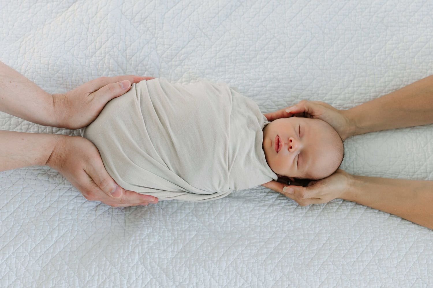 Newborn baby swaddled in white wrap lying on soft blanket while mom and dads hands gently support the infant's head and body.