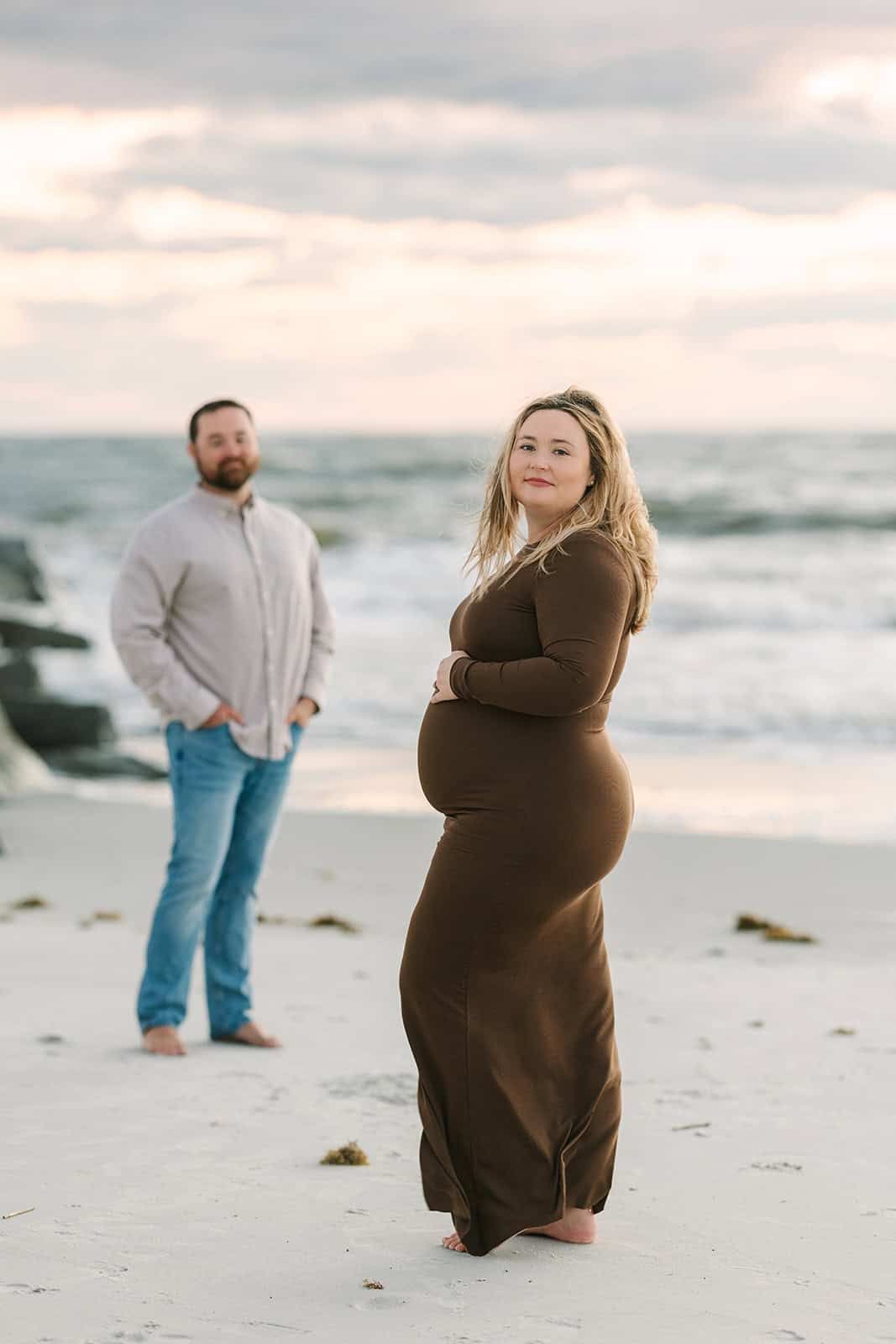 pregnant mom standing on the beach in st Augustine fl while dad looks admiringly at her