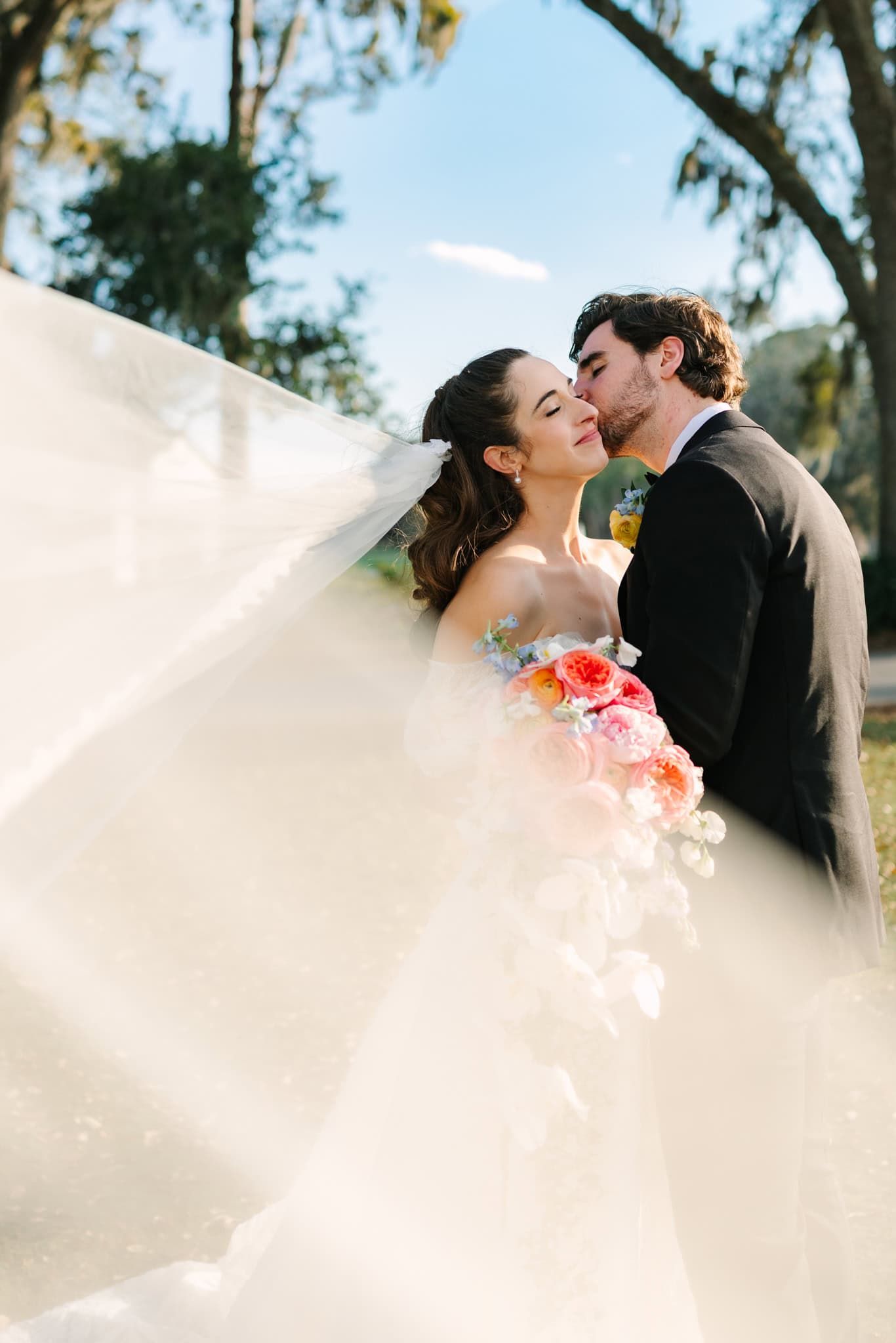 groom kissing bride on the cheek as veil blows in front of them