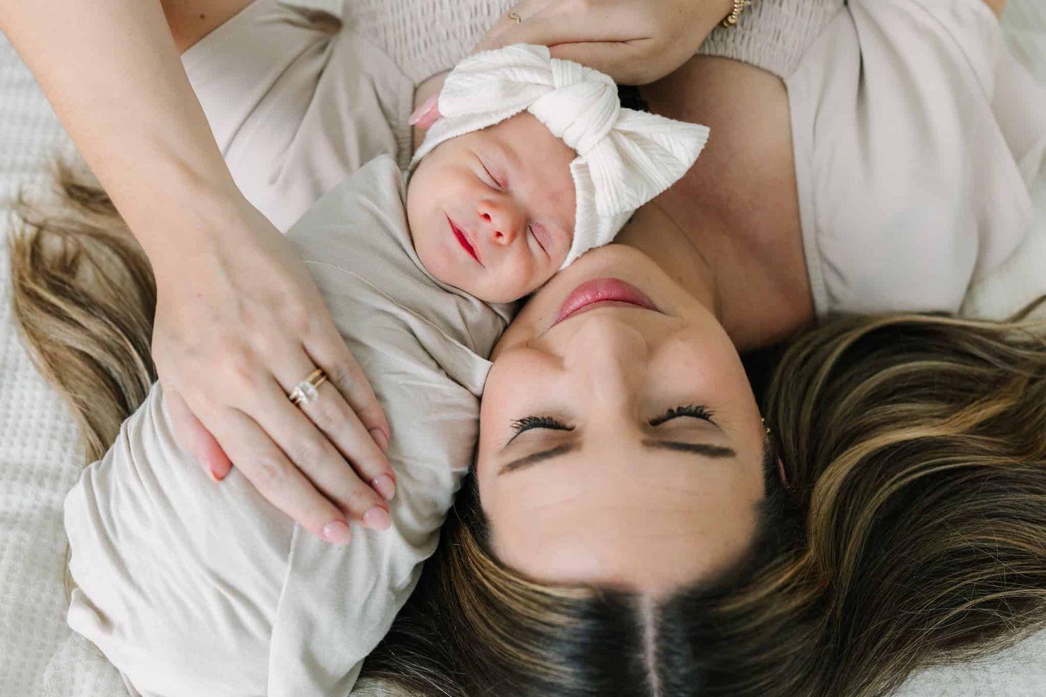 Mother and newborn baby girl lying together on a bed, with the infant wearing a white bow headband.