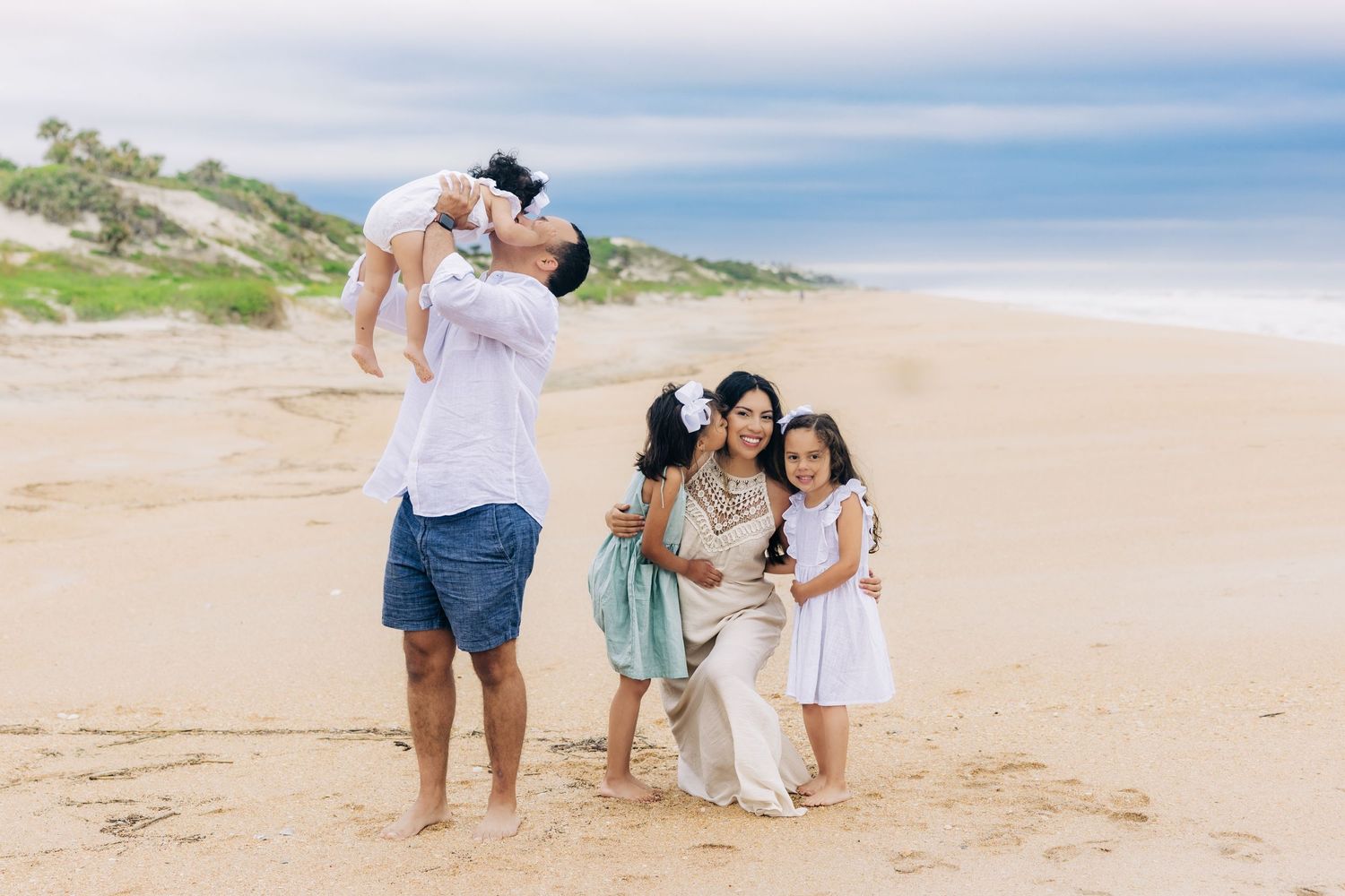 dad holding baby up in the air kissing her on jacksonville beach fl and mom kneeling down with other daughters