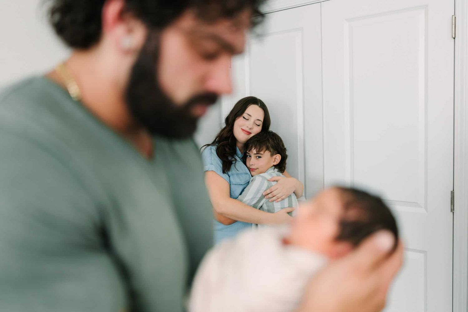 A family portrait with a bearded father holding a newborn while a mother embraces their young child against a white door.