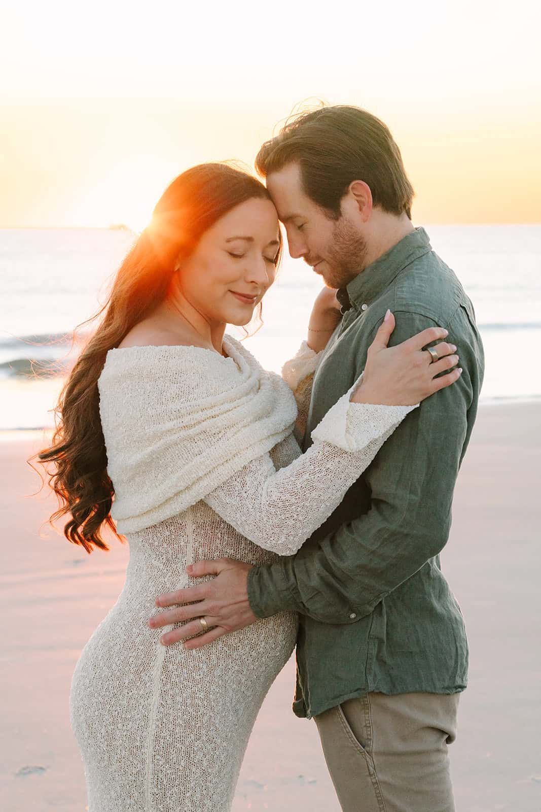 mom and dad holding each other, she is pregnant in a white dress at a Jacksonville beach fl sunrise and the sun peeking out behind her head