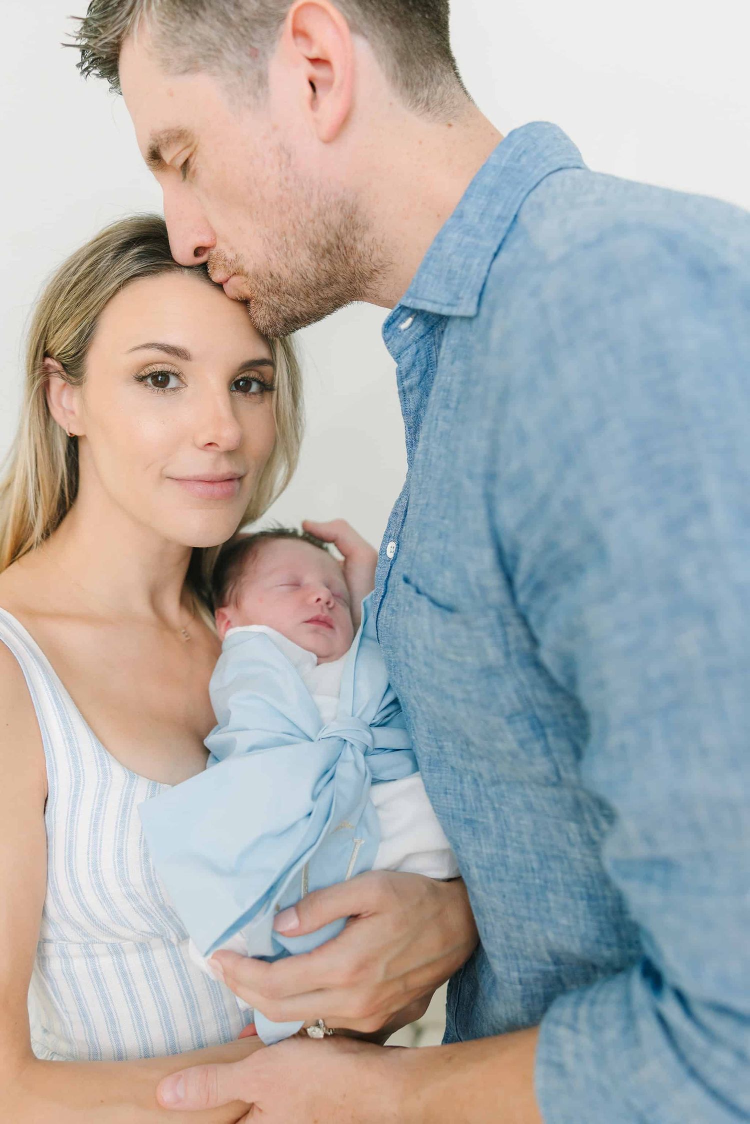 mom holding swaddled newborn baby boy with dad kissing mom on the forehead