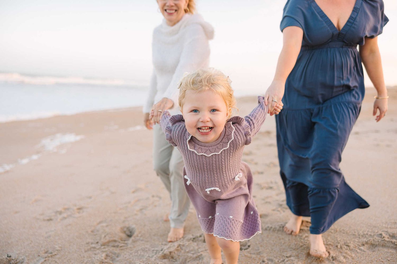 toddler girl running in the sand pulling on mom and grandmas hands to play with her