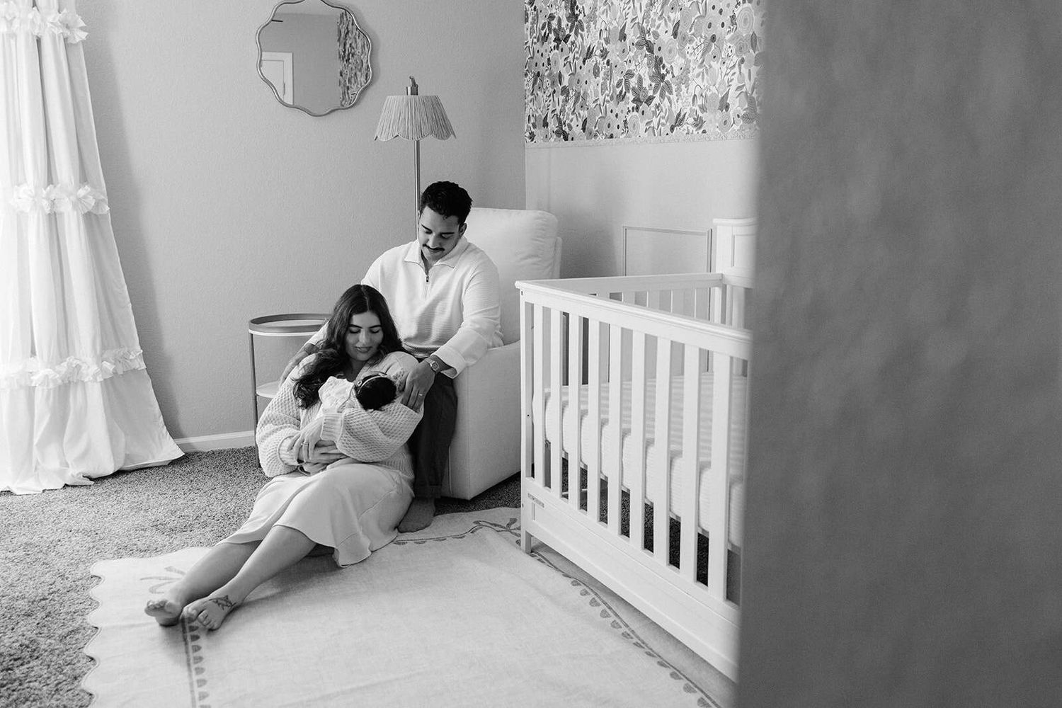 black and white peeking into the nursery of dad in glider, mom on the floor holding and snuggling newborn baby. both looking at newborn