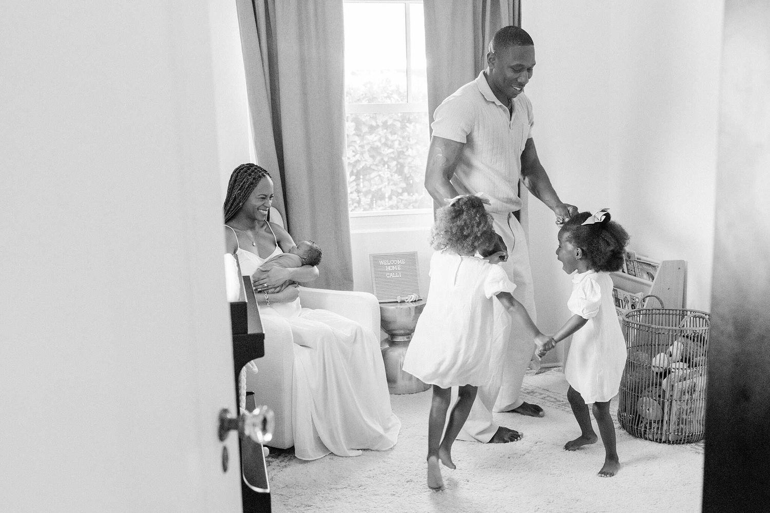 dad twirling twin toddlers while mom rocks newborn baby to sleep, peeking in from outside the nursery in black and white