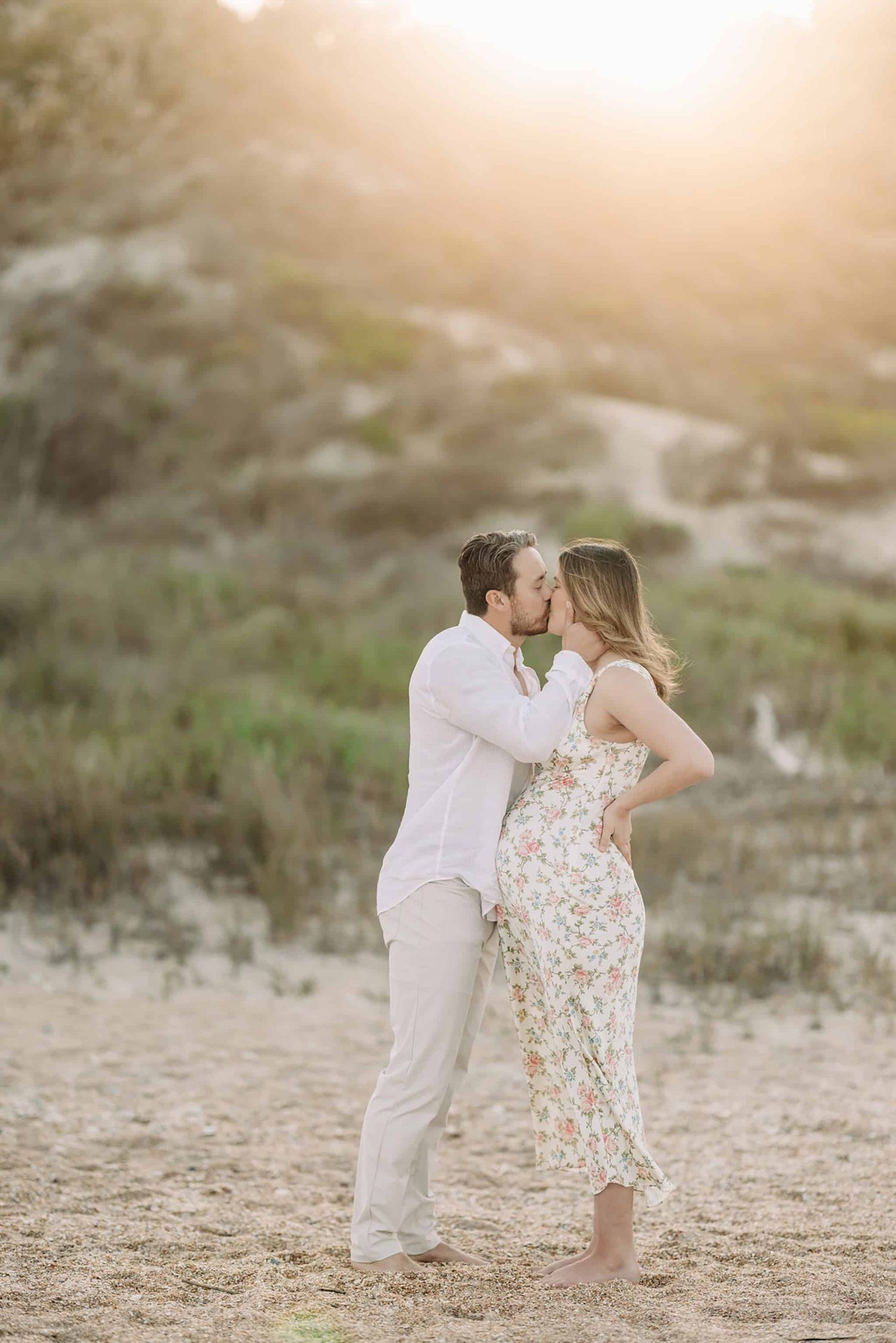 dad giving mom a big kiss on the beach with the sunset peeking over the dunes