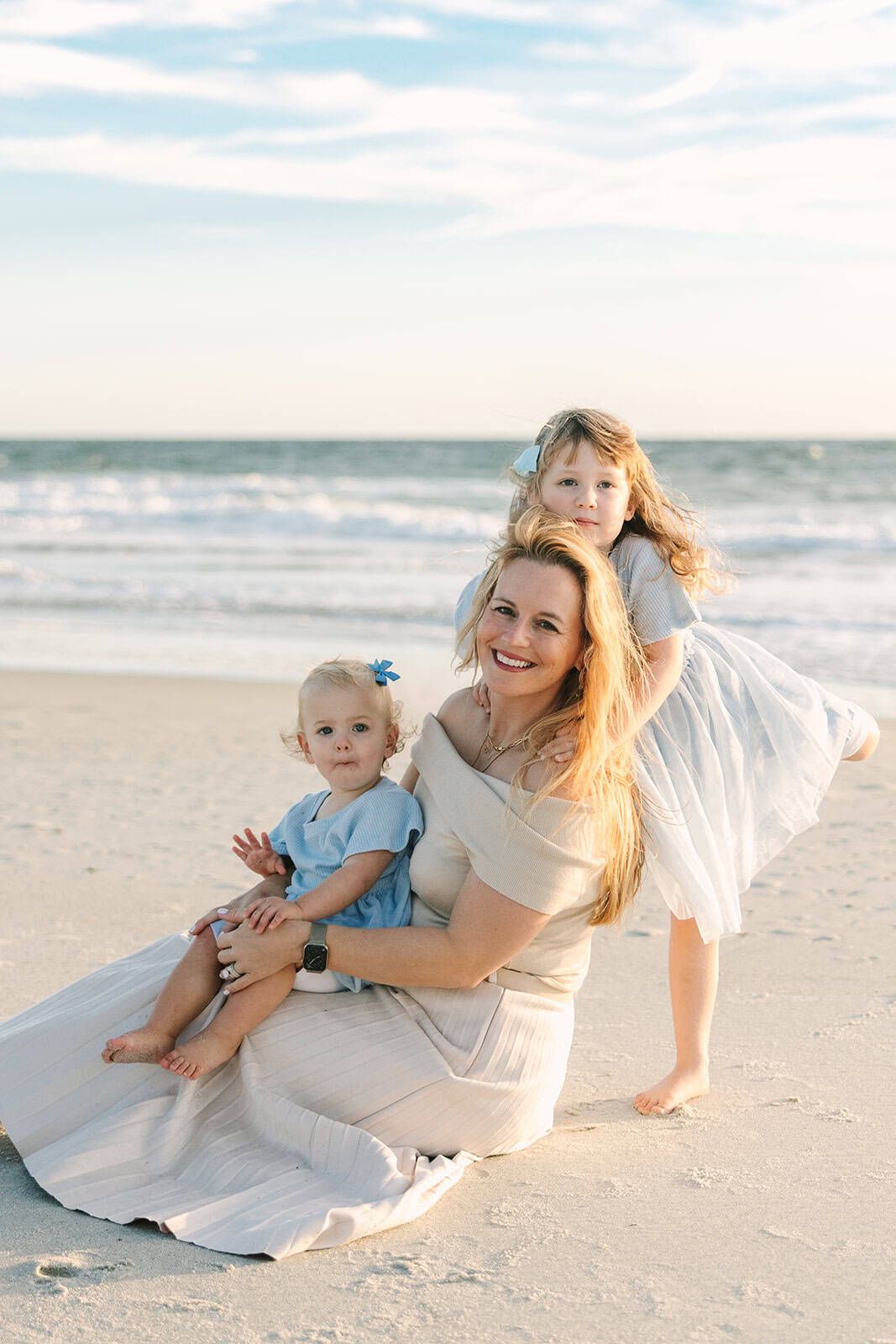 mom sitting on the beach with baby girl in her lap and older daughter hugging her from behind