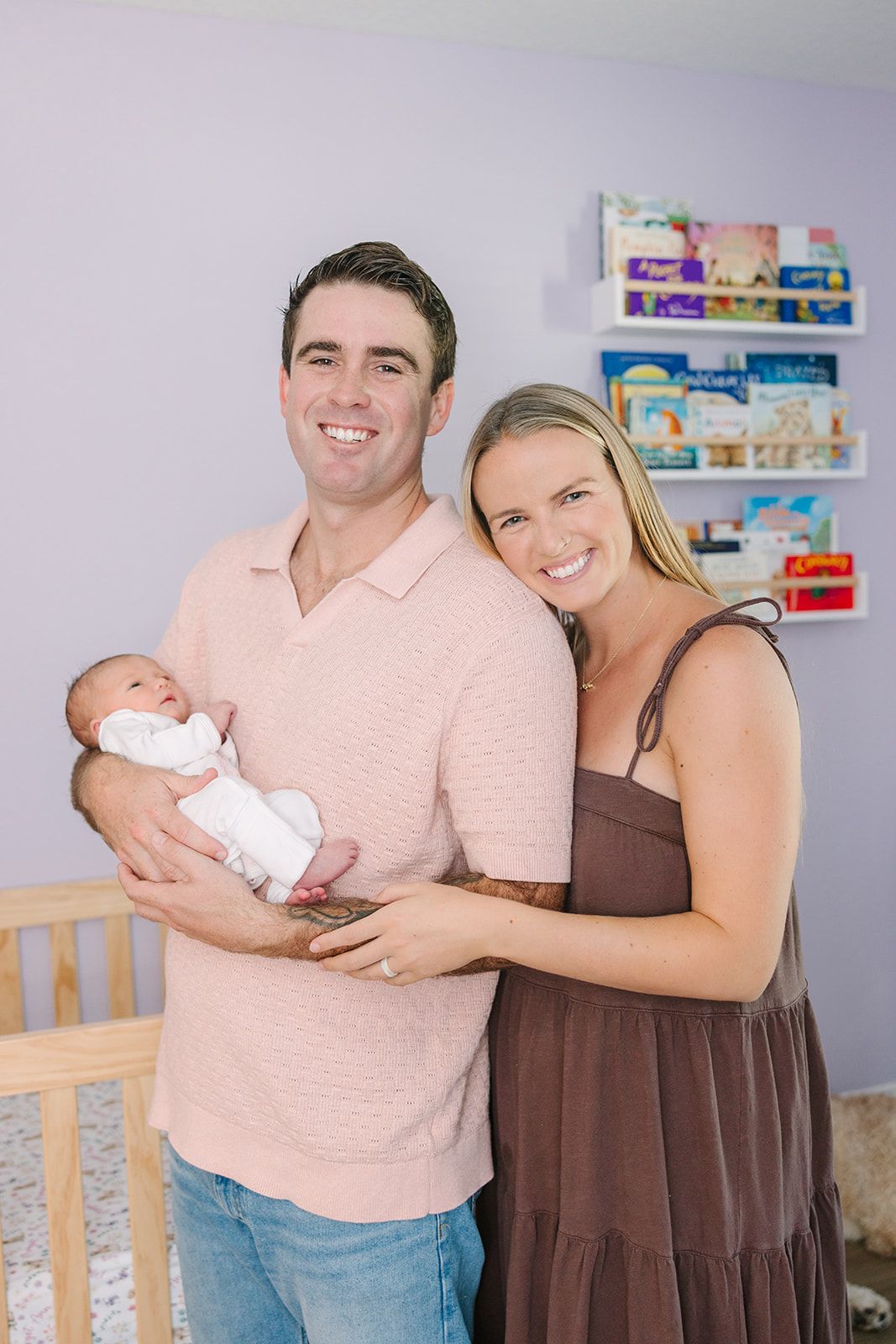 new family of dad holding baby in a pink shirt while mom hugs him from behind