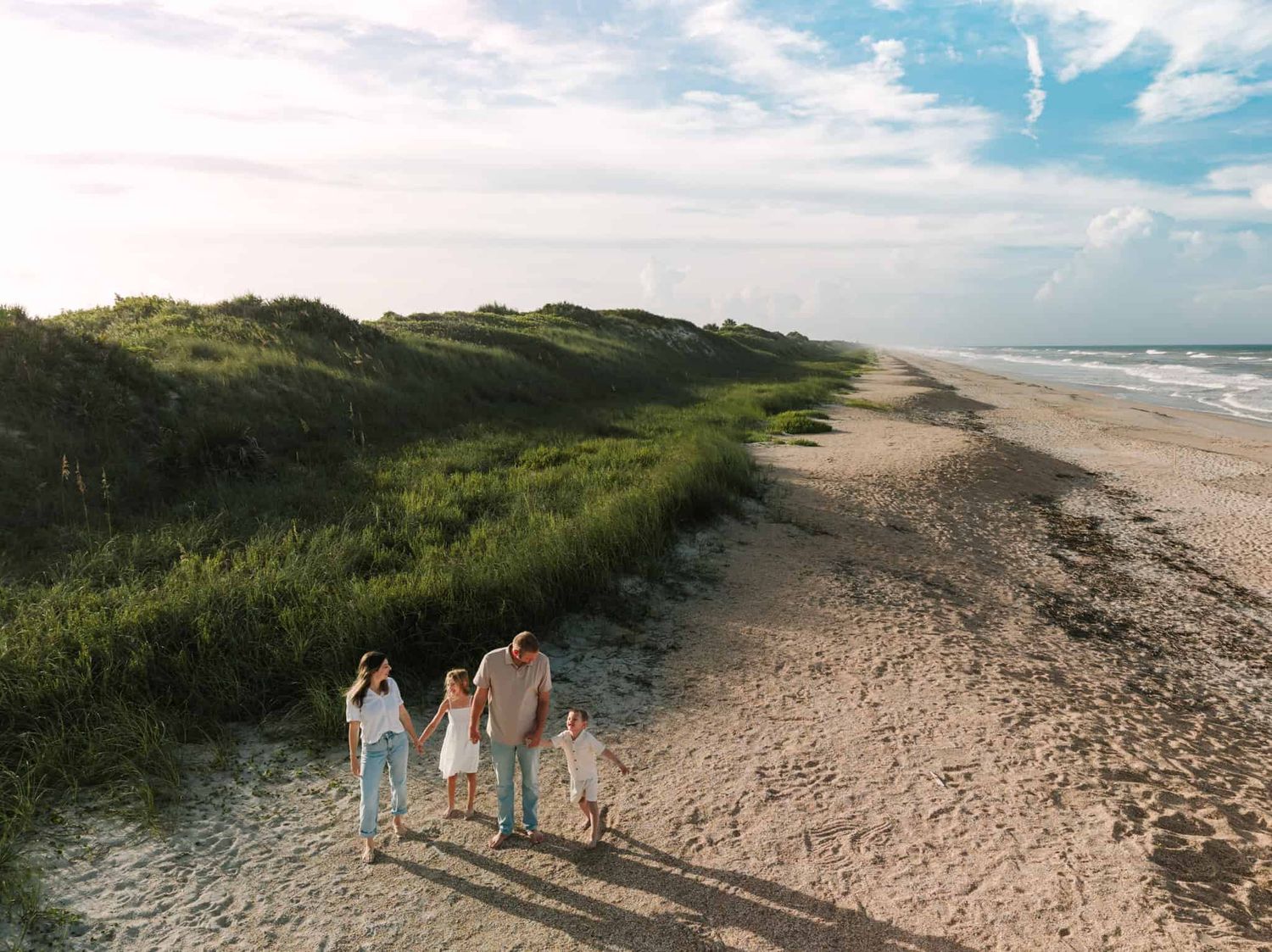 drone image Family of four walking hand-in-hand along a scenic sandy beach path beside lush green dunes at sunset.