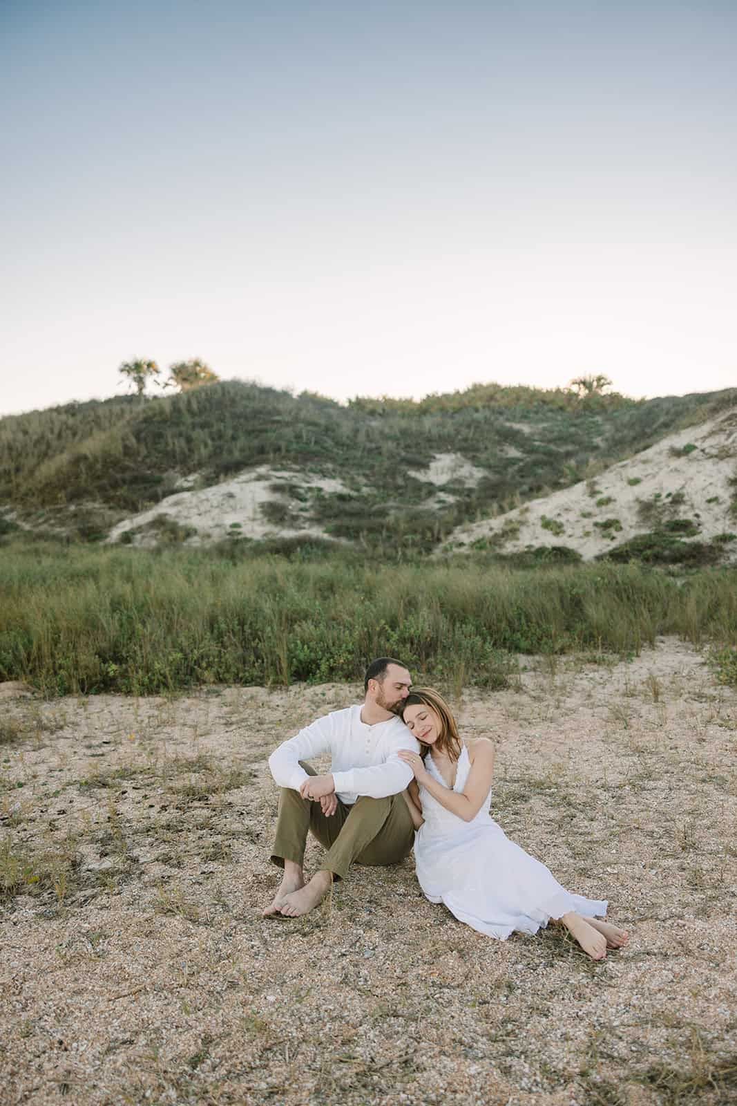 A couple sits together on sandy ground near green hills, the woman in white dress leaning on mans shoulder at sunrise