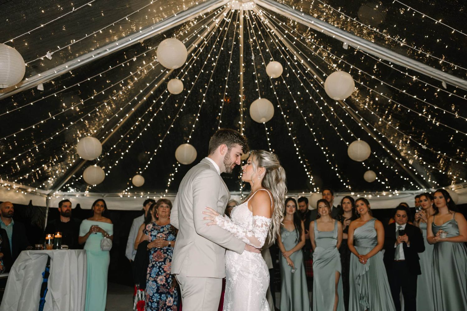 Couple shares first dance under fairy lights and paper lanterns, surrounded by bridesmaids in sage green at wedding reception.