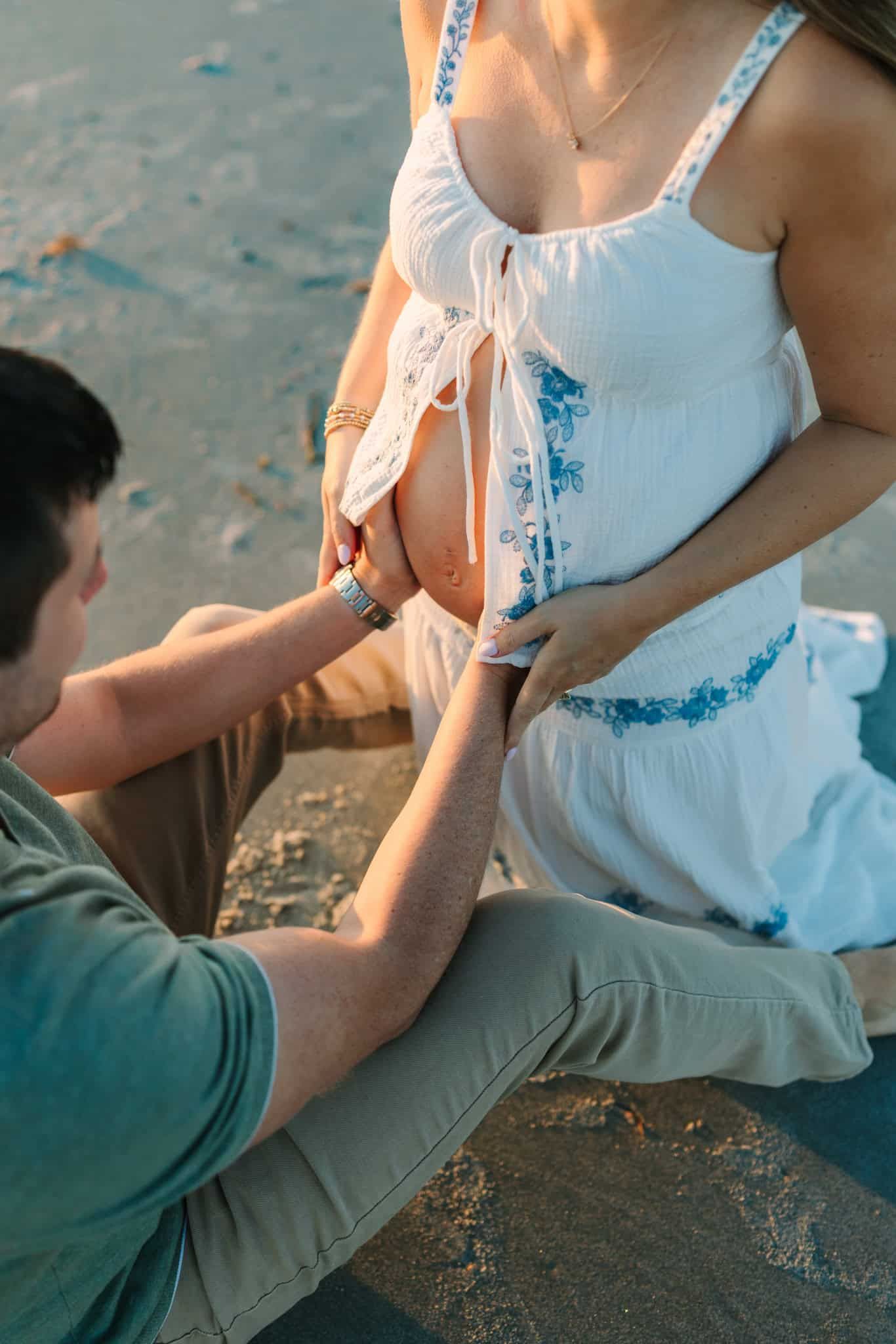 A husband tenderly touches a pregnant woman's belly outdoors on a beach during golden hour.