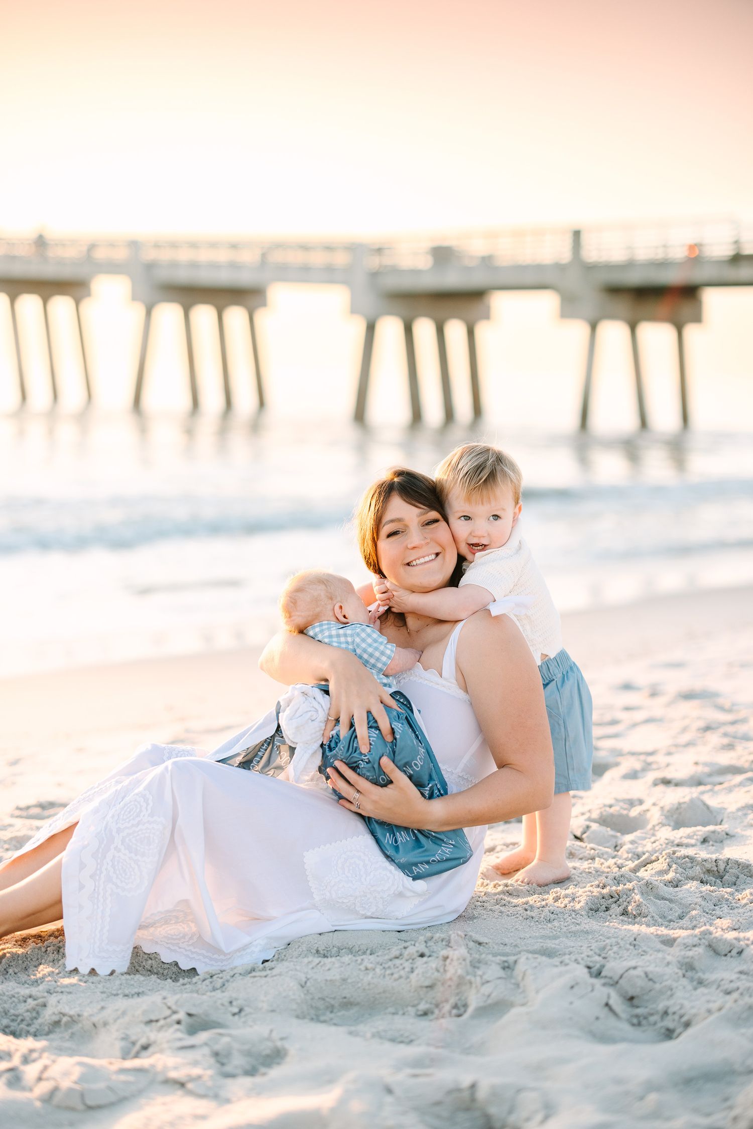 Caitlin sitting in the sand  holding baby Nolan with Bennett behind her giving her a big hug