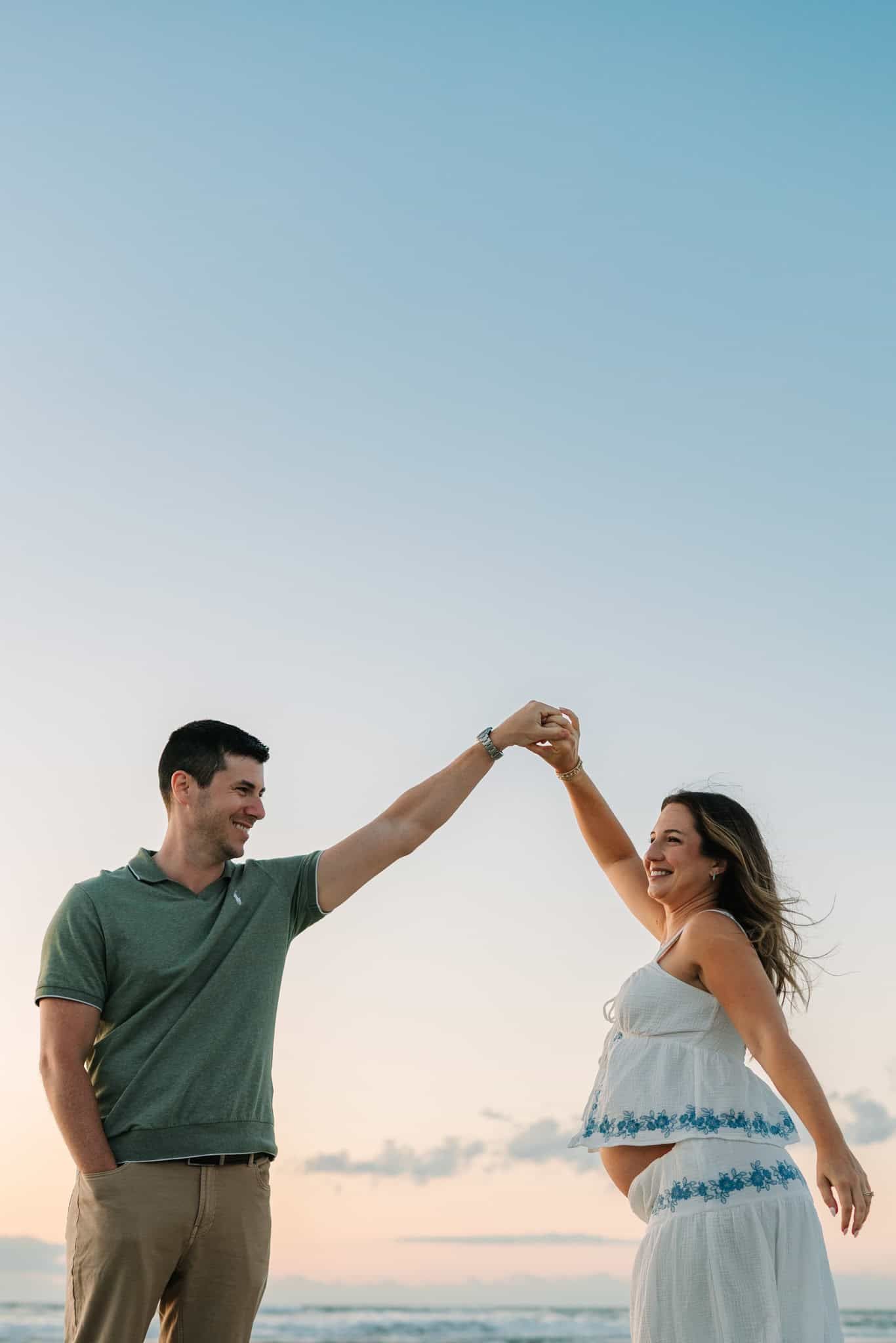 A happy couple dances on a beach at sunset, man in green polo spinning woman in white blue dress.