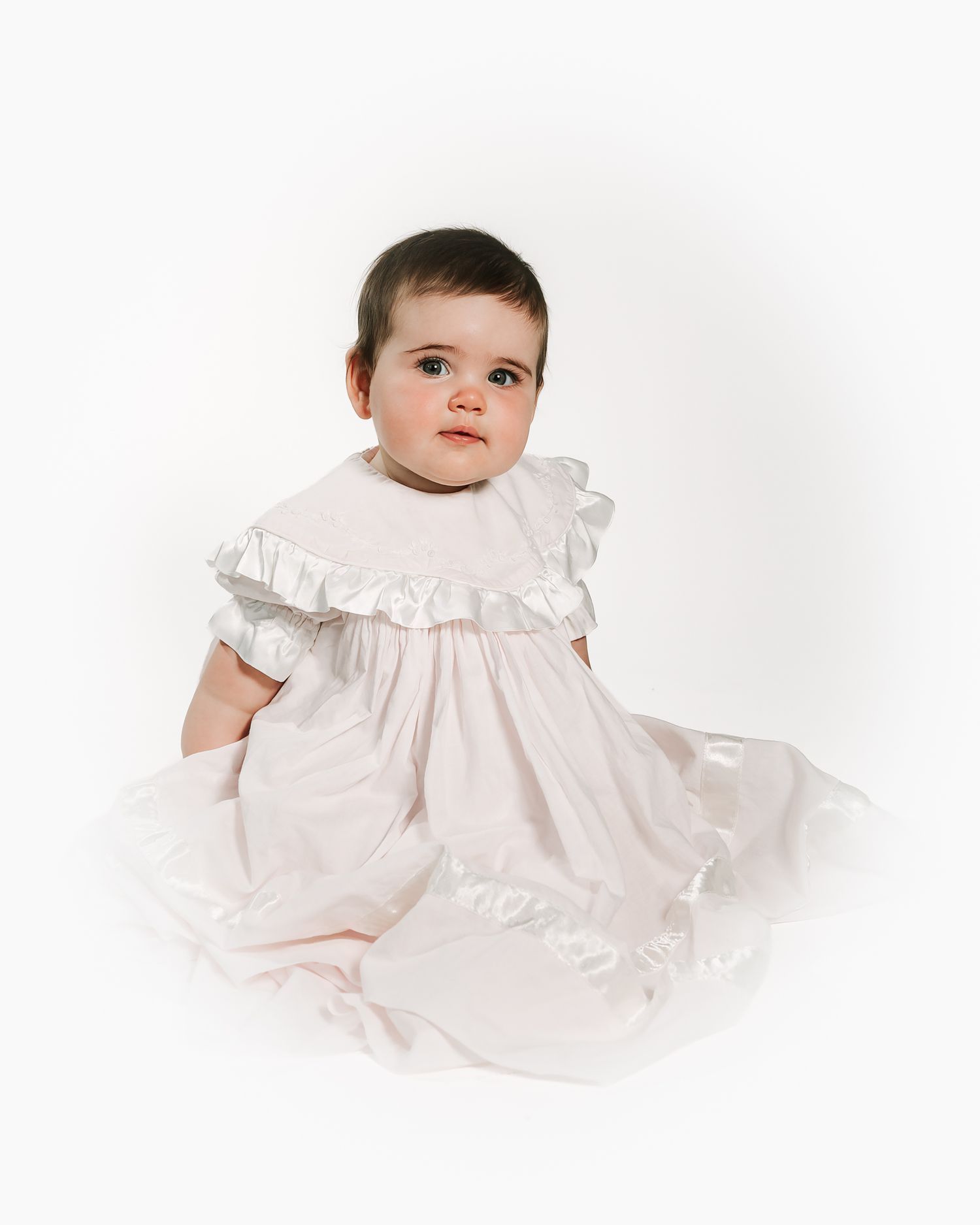 Baby girl in a white ruffled dress with satin trim sits posed against a white background.