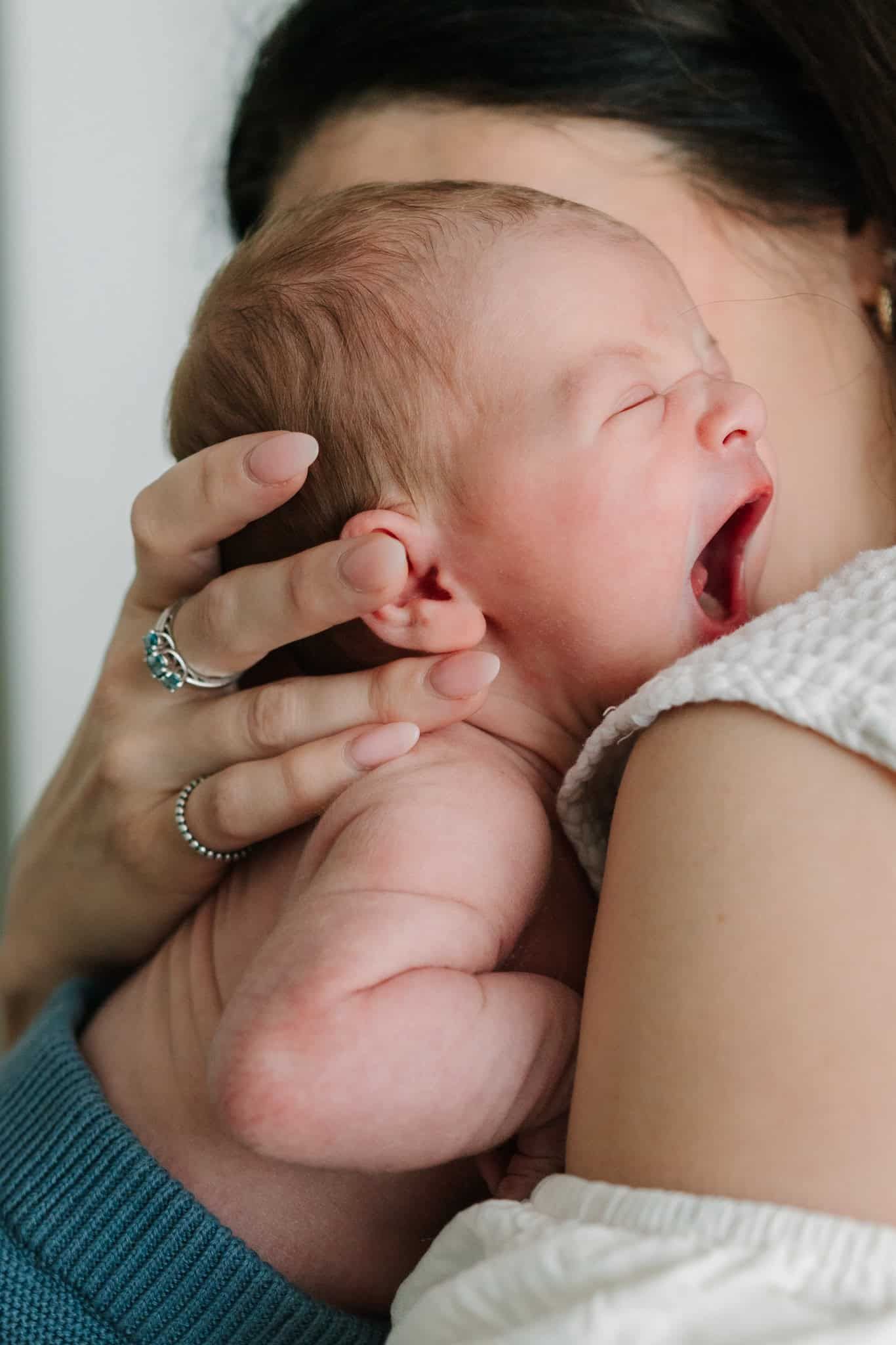 A newborn baby yawning while being cradled against a mother's chest, her hand gently supporting the infant's head.