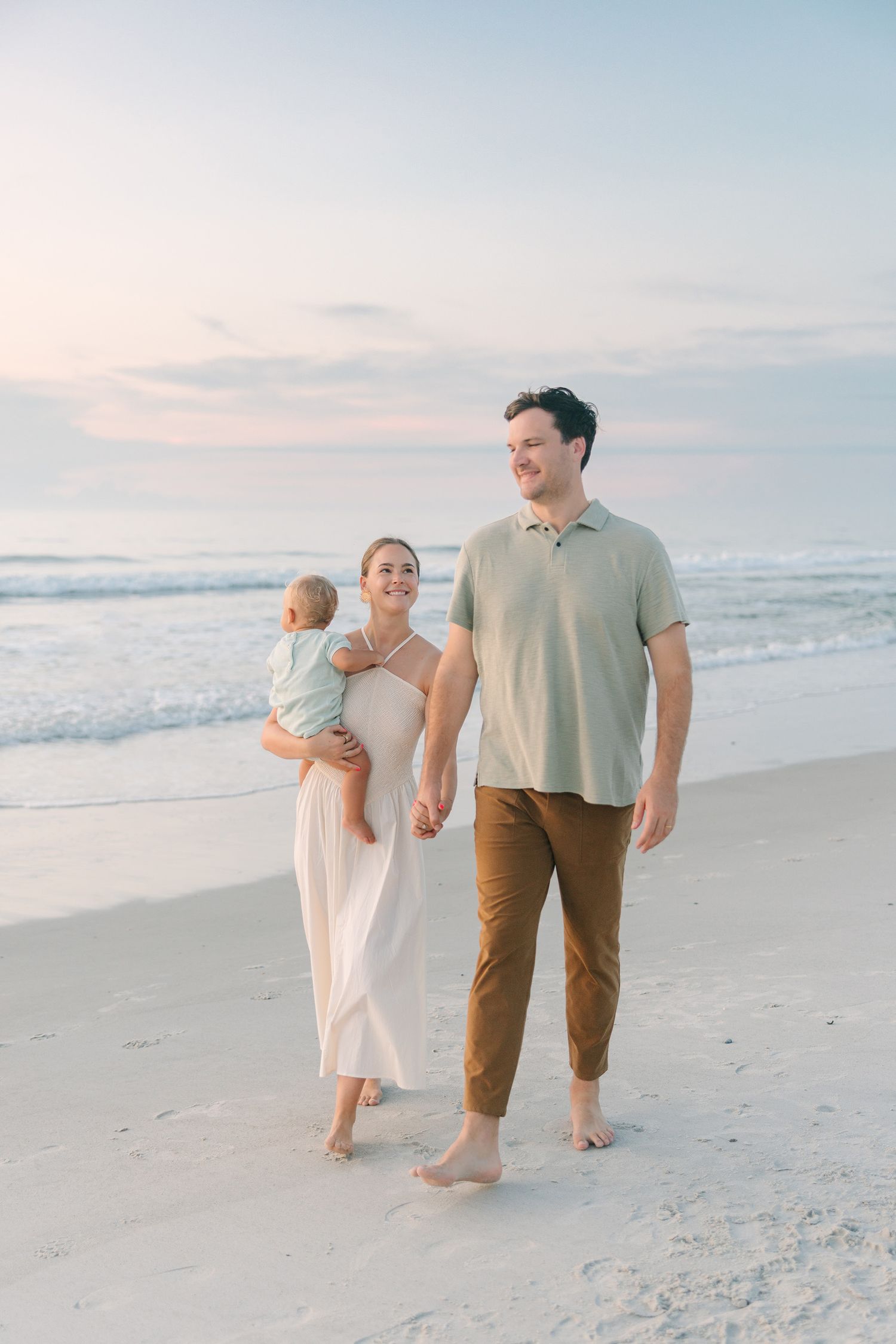 mom and dad holding hands with mom holding one year old son walking down the beach