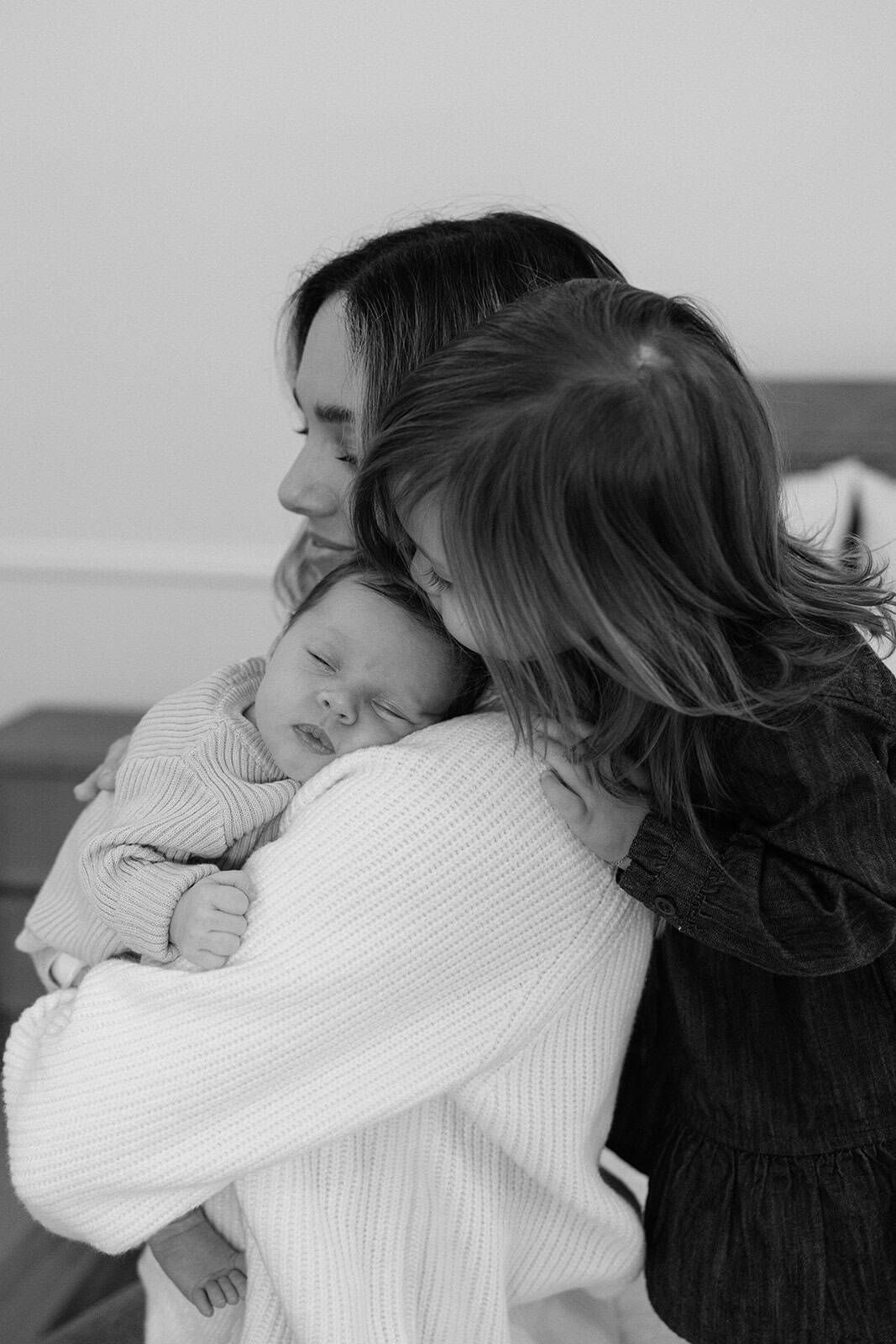 black and white photo of mom snuggling with newborn while older sister leaning over moms shoulder giving baby a kiss