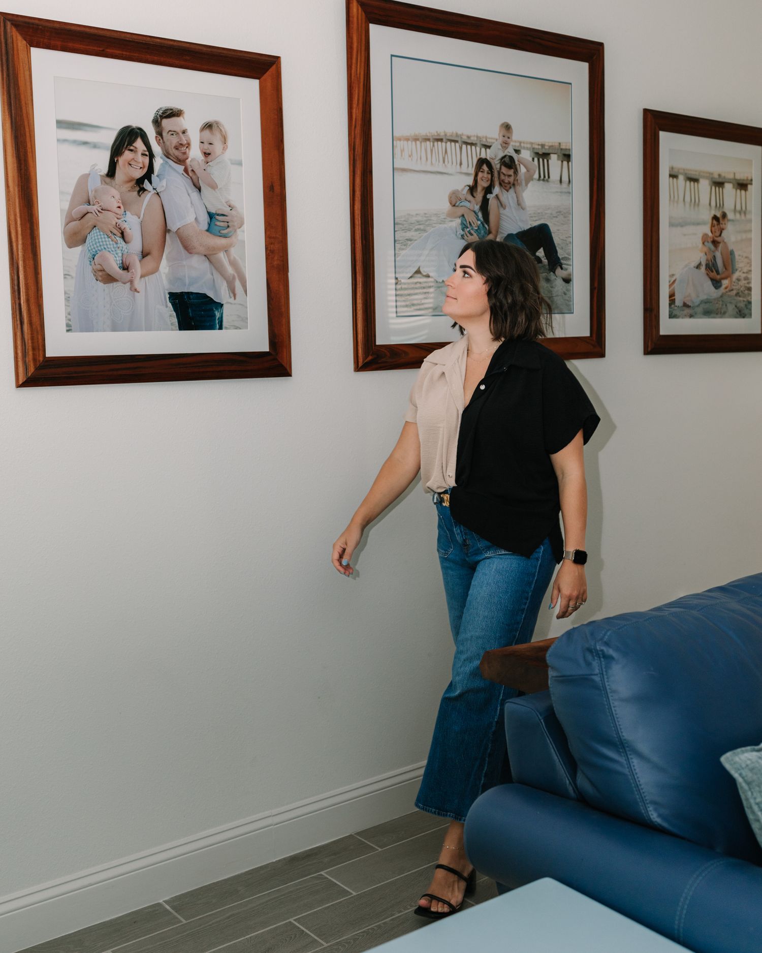Woman in black top and jeans admiring framed family portrait photos displayed on a white wall above a blue sofa.