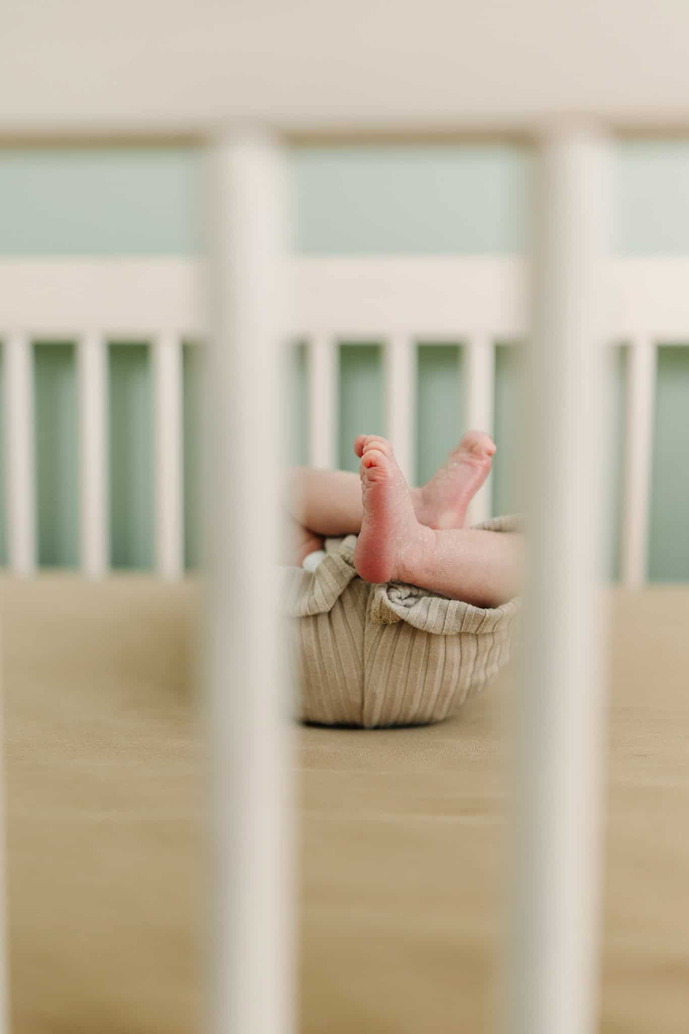 Newborn baby feet peeking out of a white knit basket inside a white wooden crib.