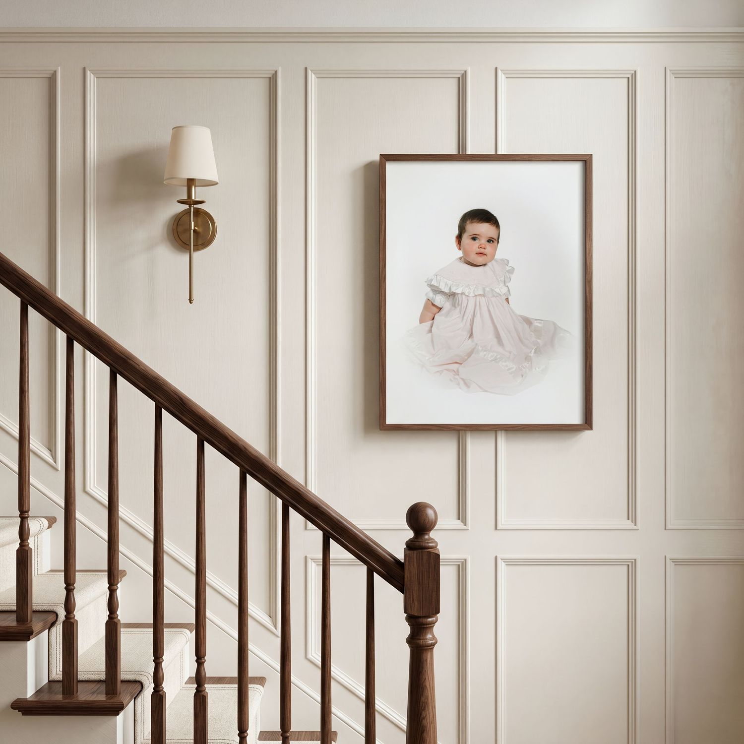 Framed baby portrait in white dress displayed on cream wainscoting wall above staircase with brass sconce.